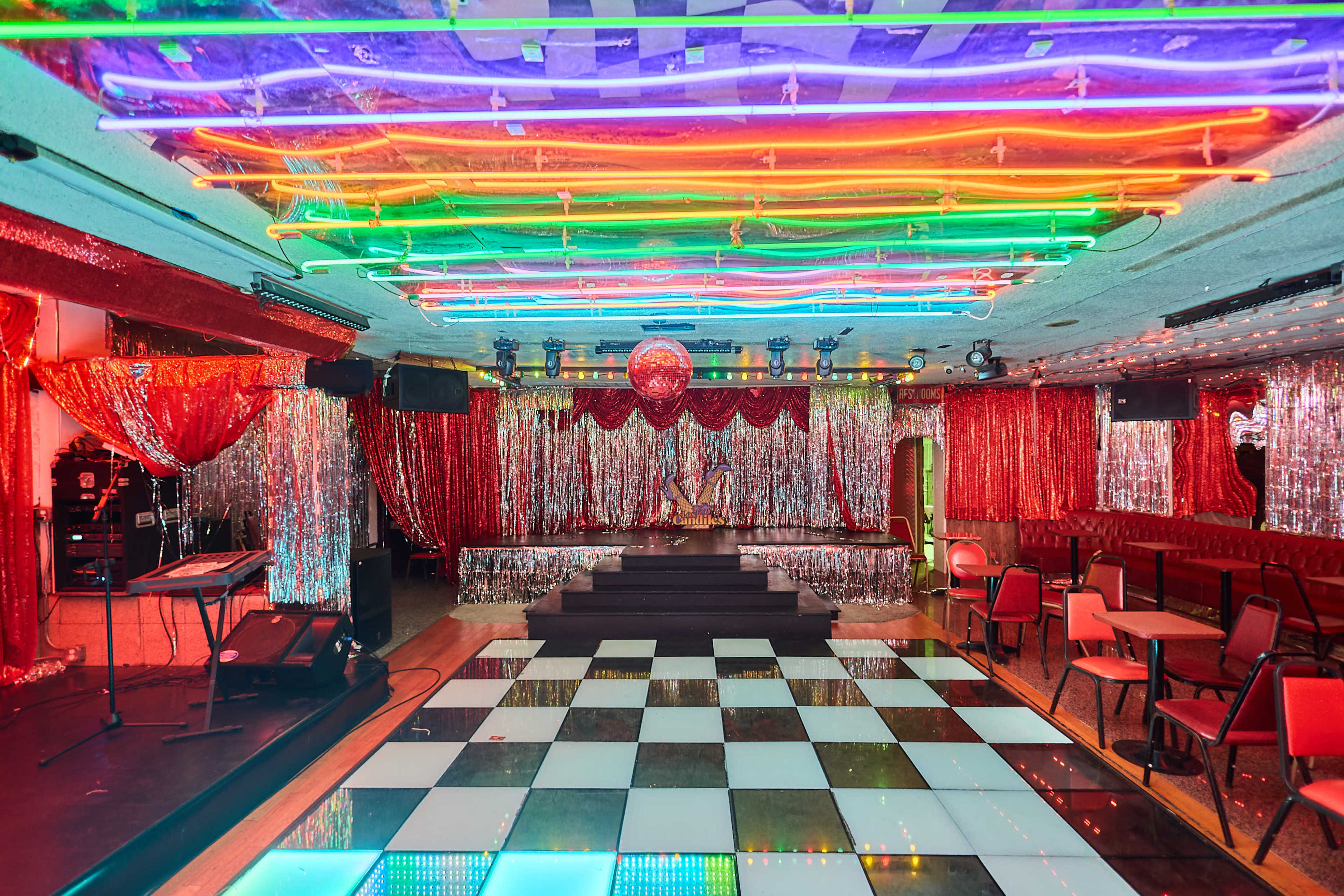 The image shows a vibrant nightclub interior with a checkered dance floor, colorful neon lights, glittering decorations, and a stage set up for performances.