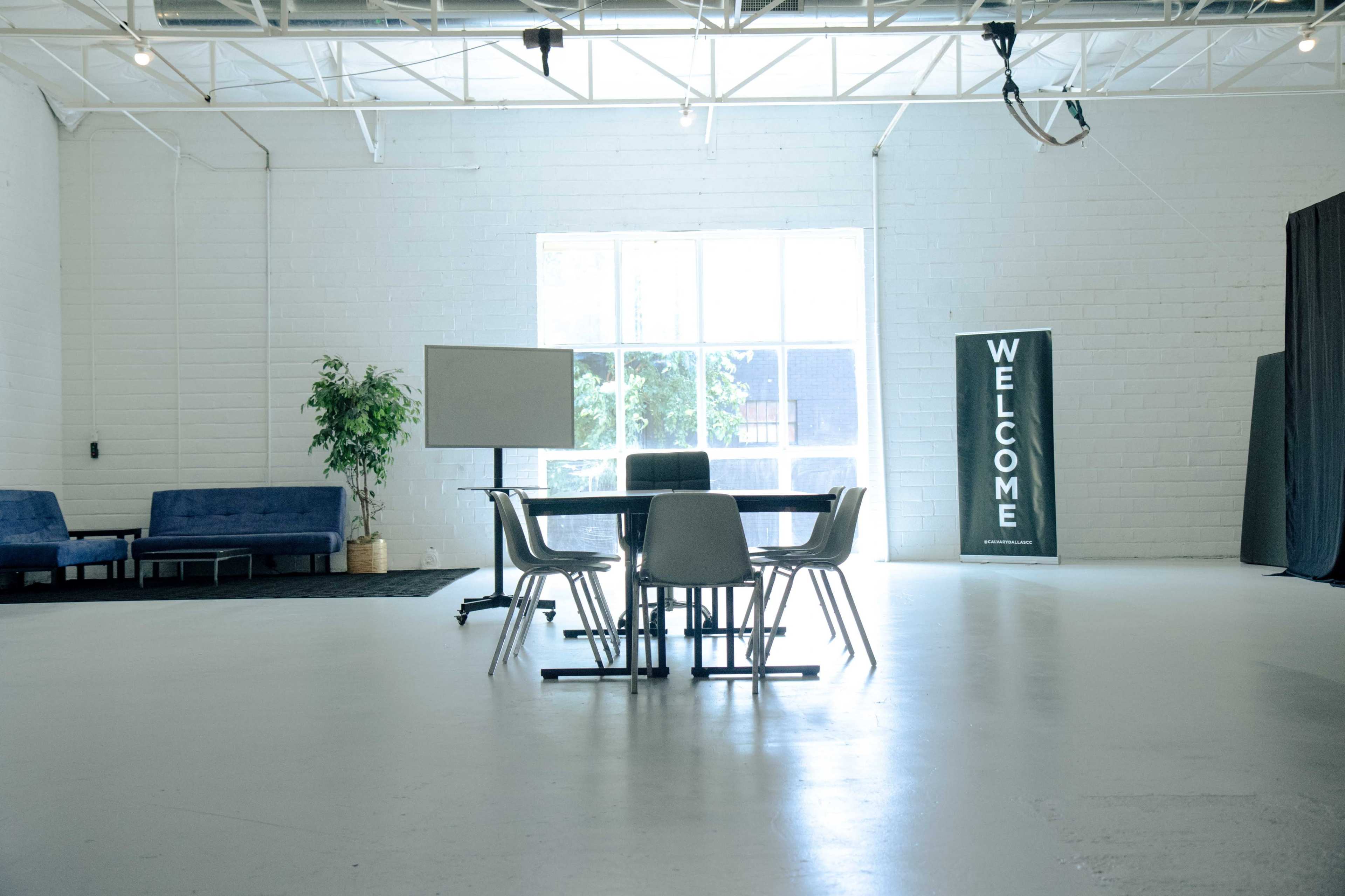 A bright, spacious room features a round table surrounded by chairs, with a large window and a "WELCOME" sign on one side.