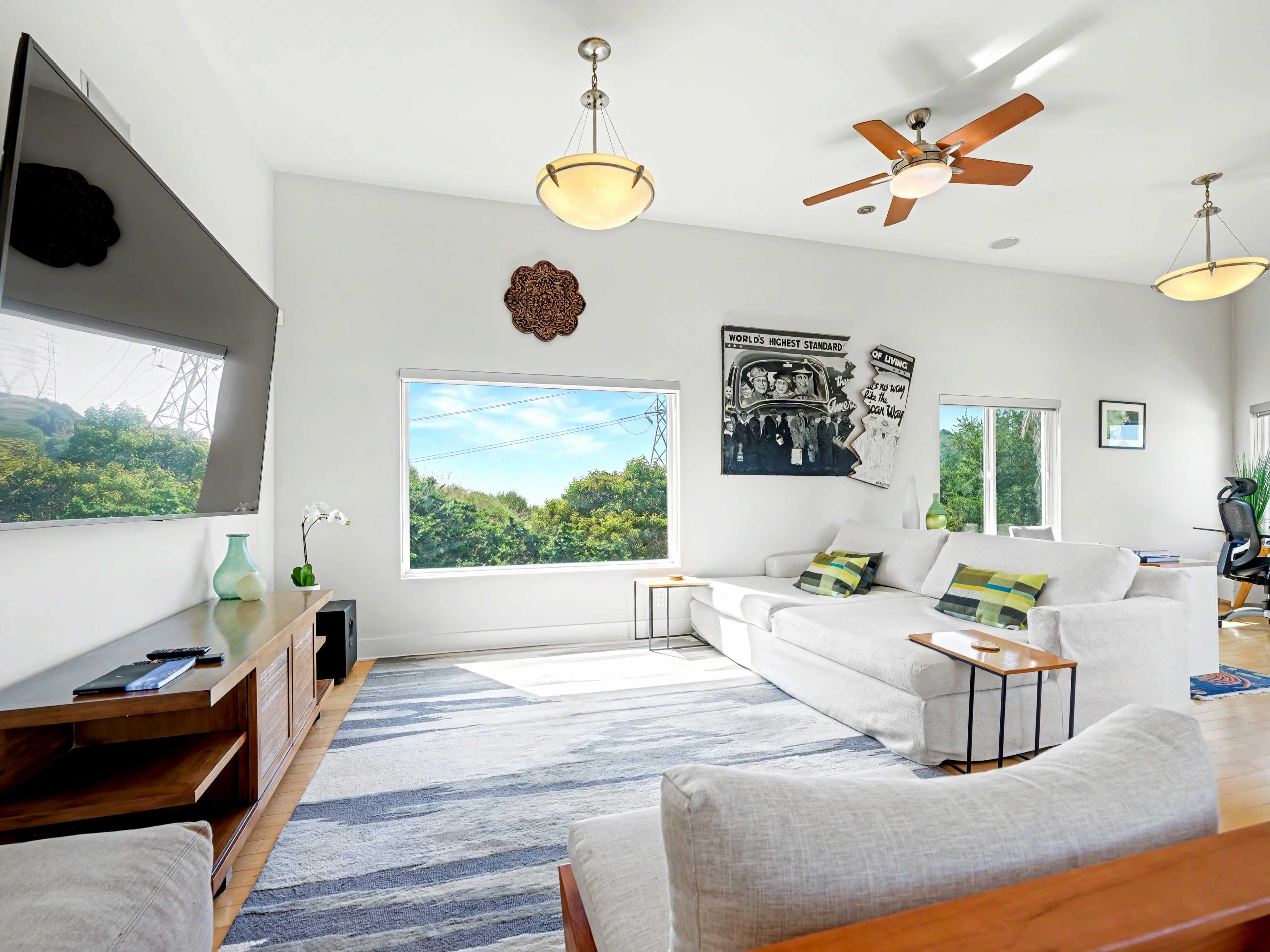 The image shows a bright living room with a large window, light-colored sofas, a wooden coffee table, and a wall-mounted television.