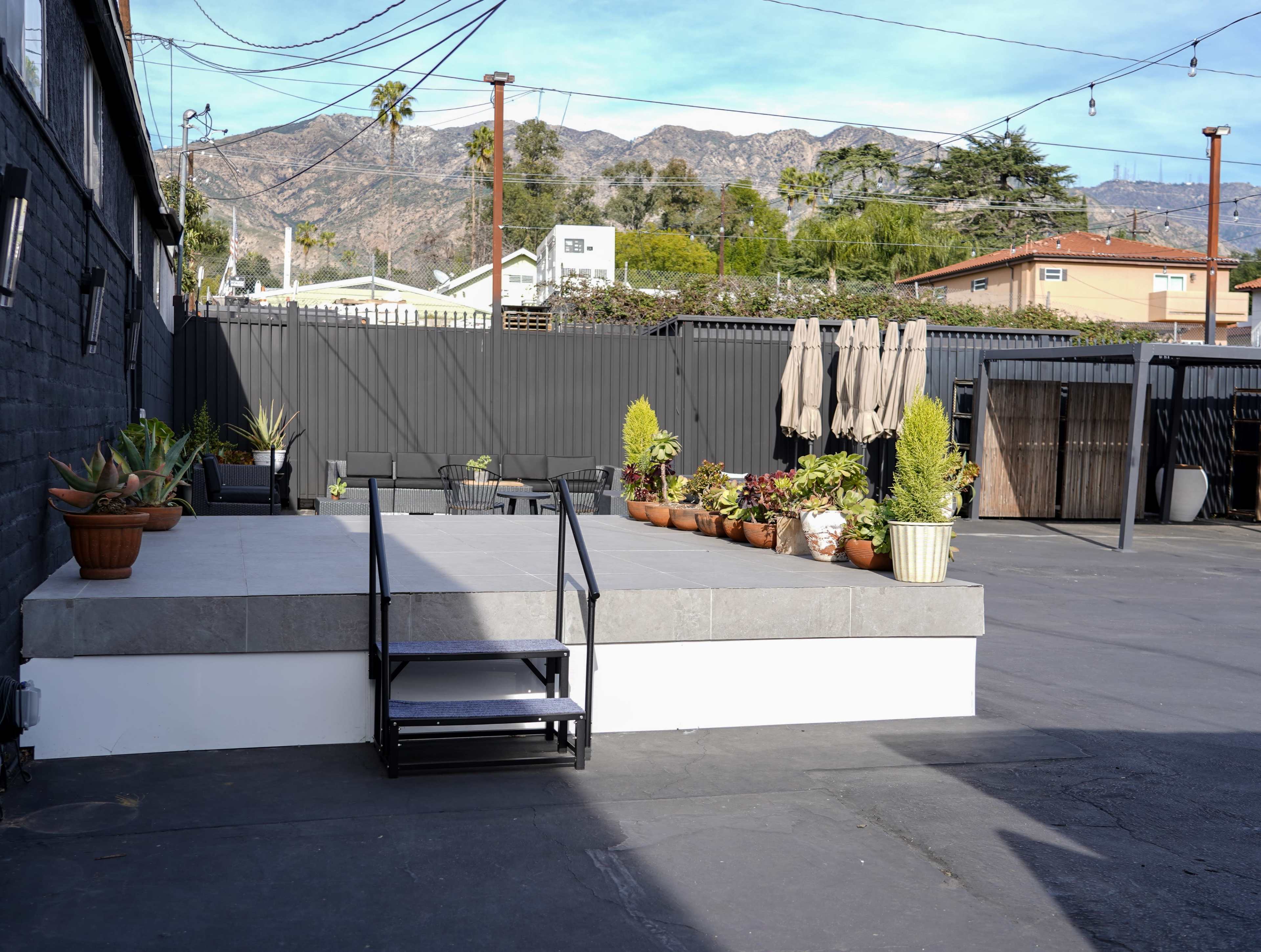 The image shows a patio area with potted plants, a set of stairs, and several umbrellas, against a backdrop of mountains.