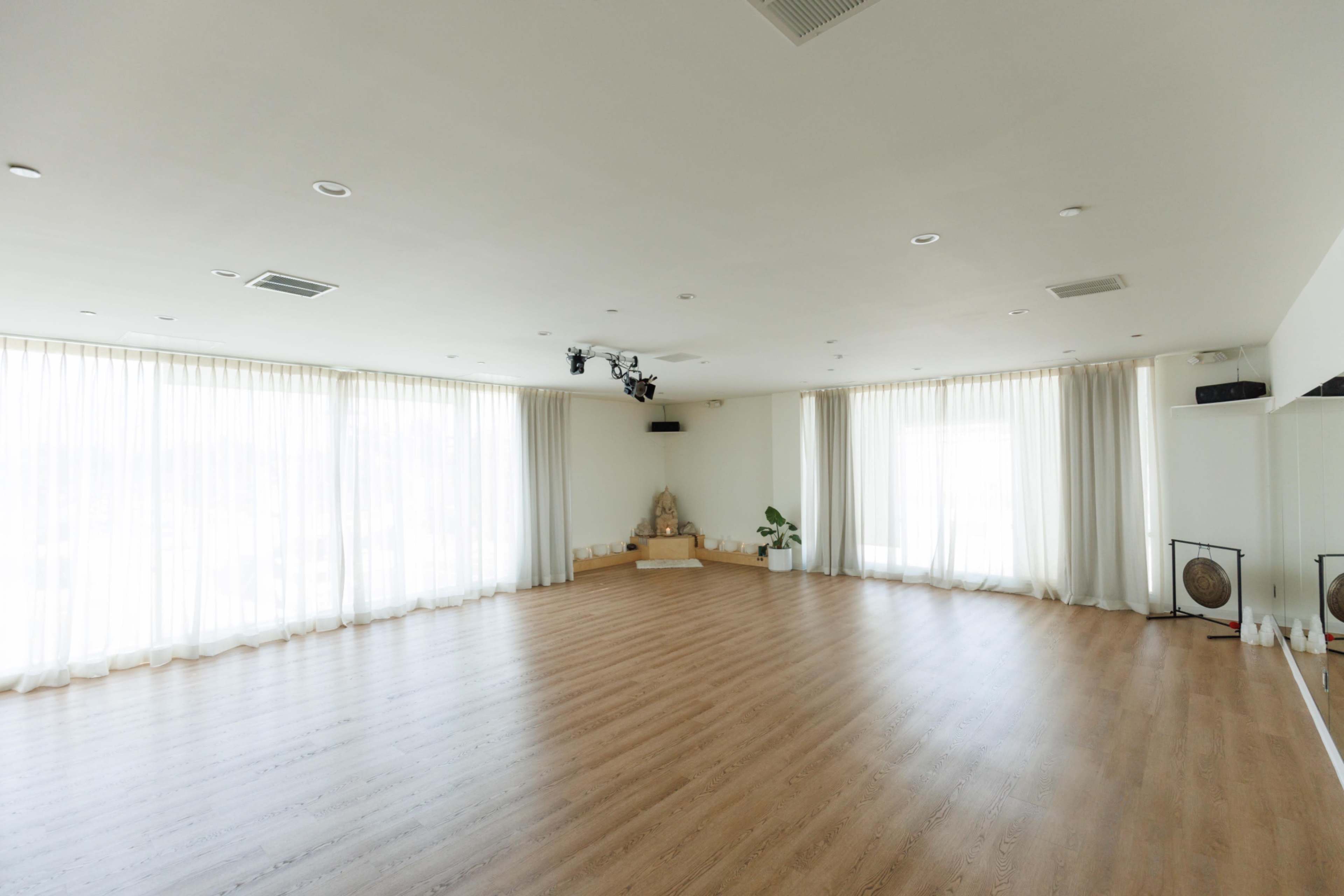 The image shows a spacious, empty room with large windows covered by sheer curtains and wooden flooring.