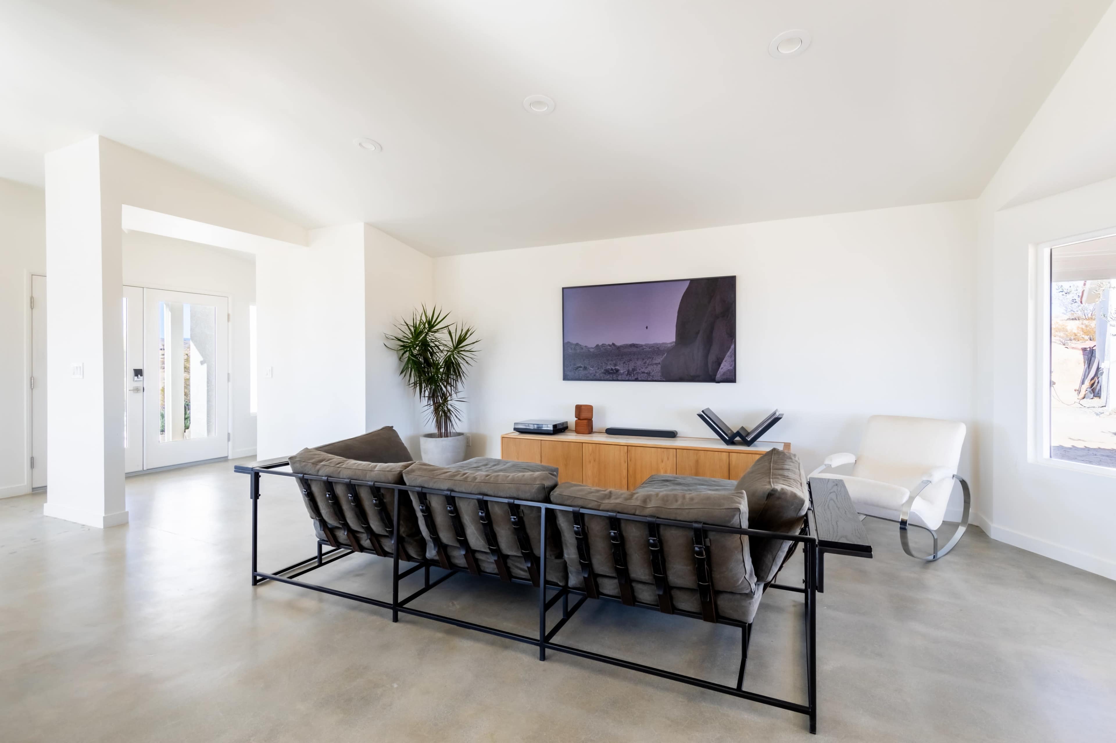 A modern living room features a large television mounted on a white wall, flanked by a wooden media console, with a gray sofa and a minimalist armchair in the foreground.