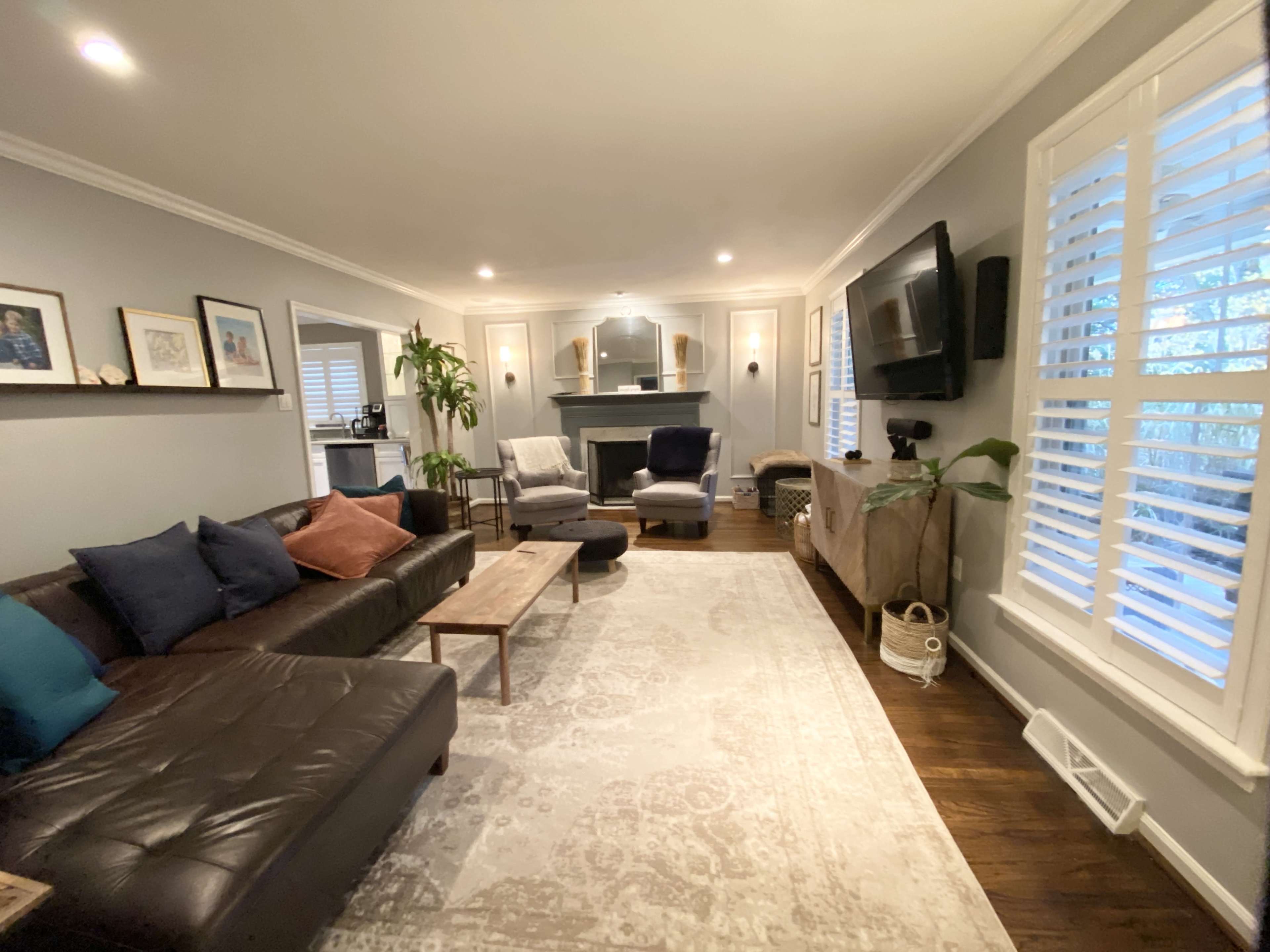 The living room features a dark leather sofa, a wooden coffee table, and a cozy seating area with two armchairs, framed artwork on the walls, and large windows with plantation shutters.