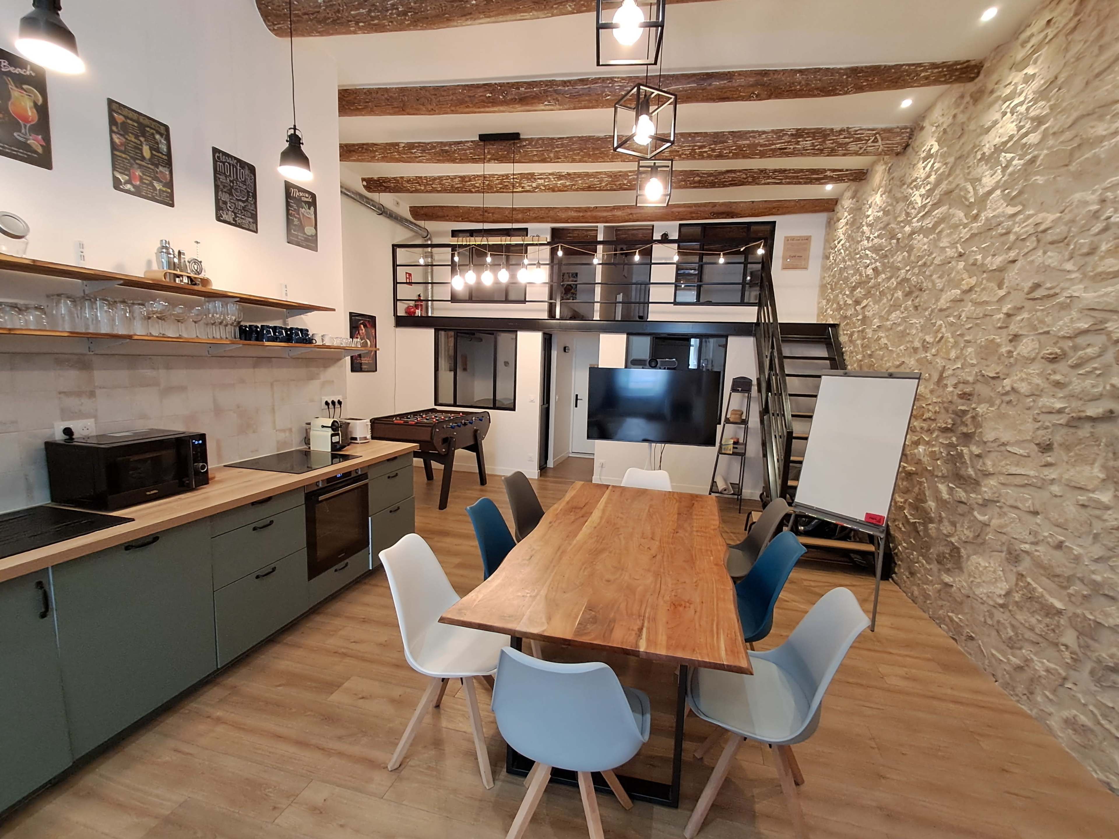 The image shows a modern open kitchen and dining area featuring a wooden table surrounded by colorful chairs, a foosball table, and a staircase leading to a loft.