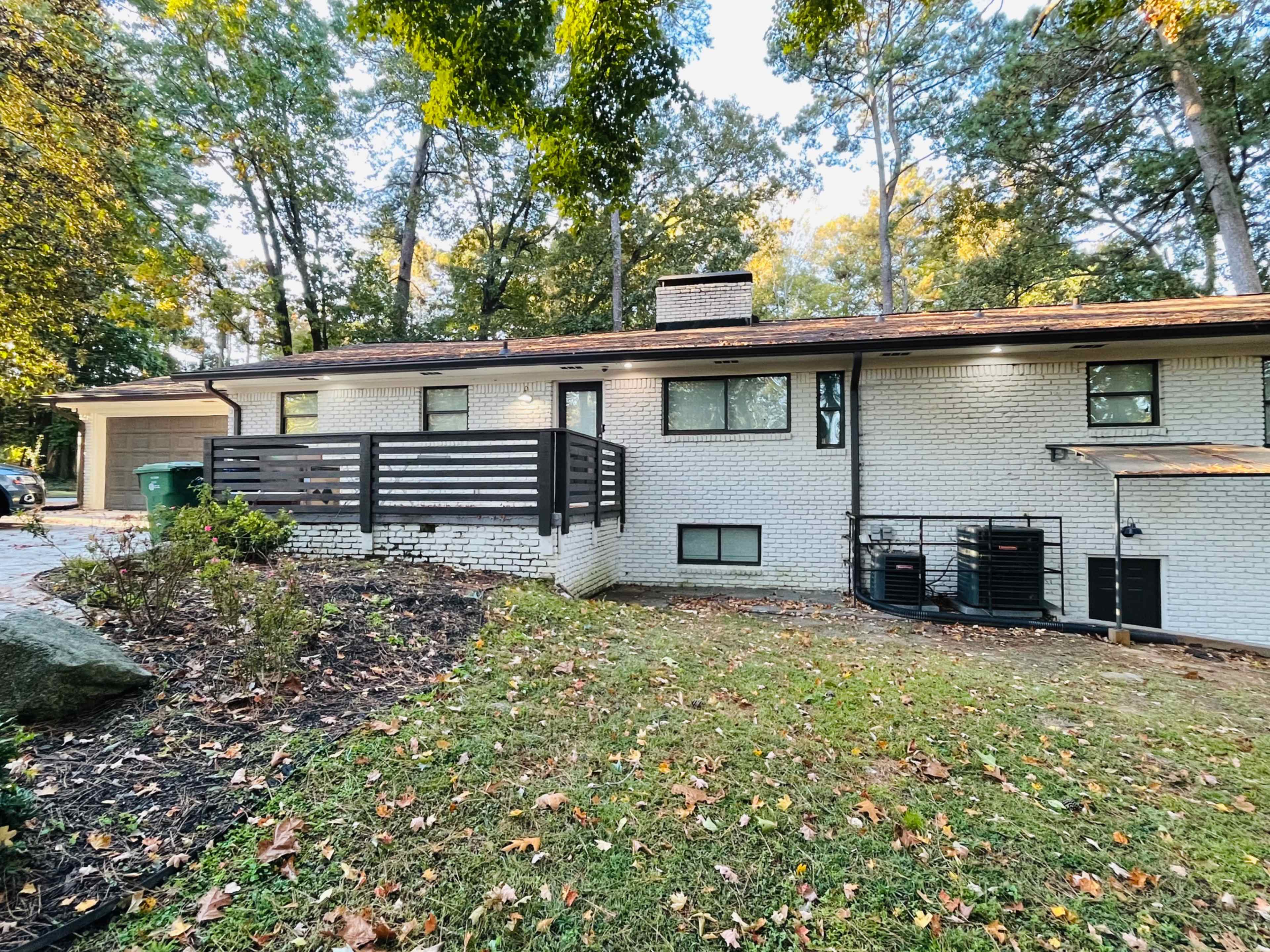The image shows a single-story house with a brick exterior, a wooden deck, and surrounded by trees and landscaping.