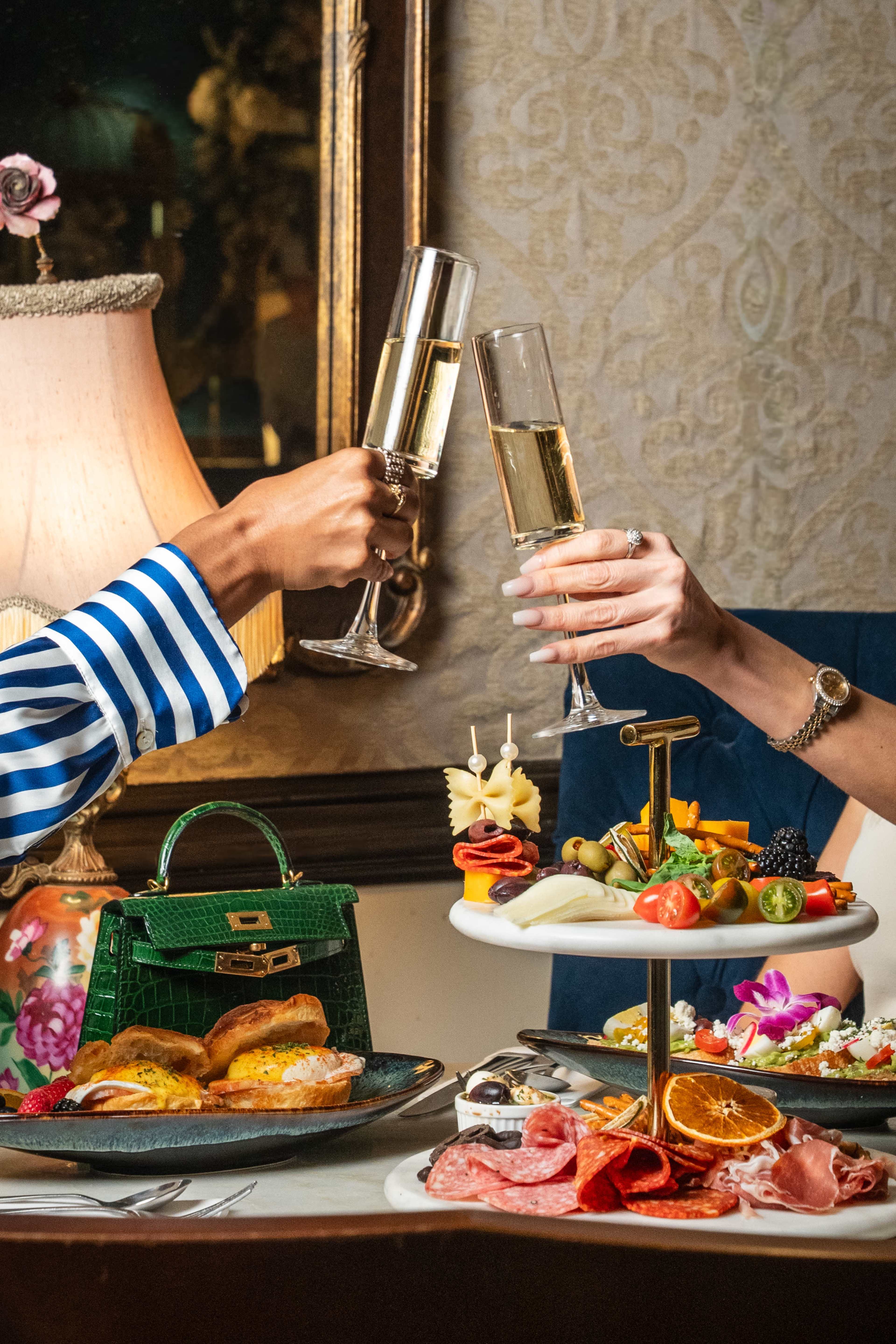 Two hands hold champagne flutes in a toast above a table adorned with an assortment of appetizers and a stylish handbag.