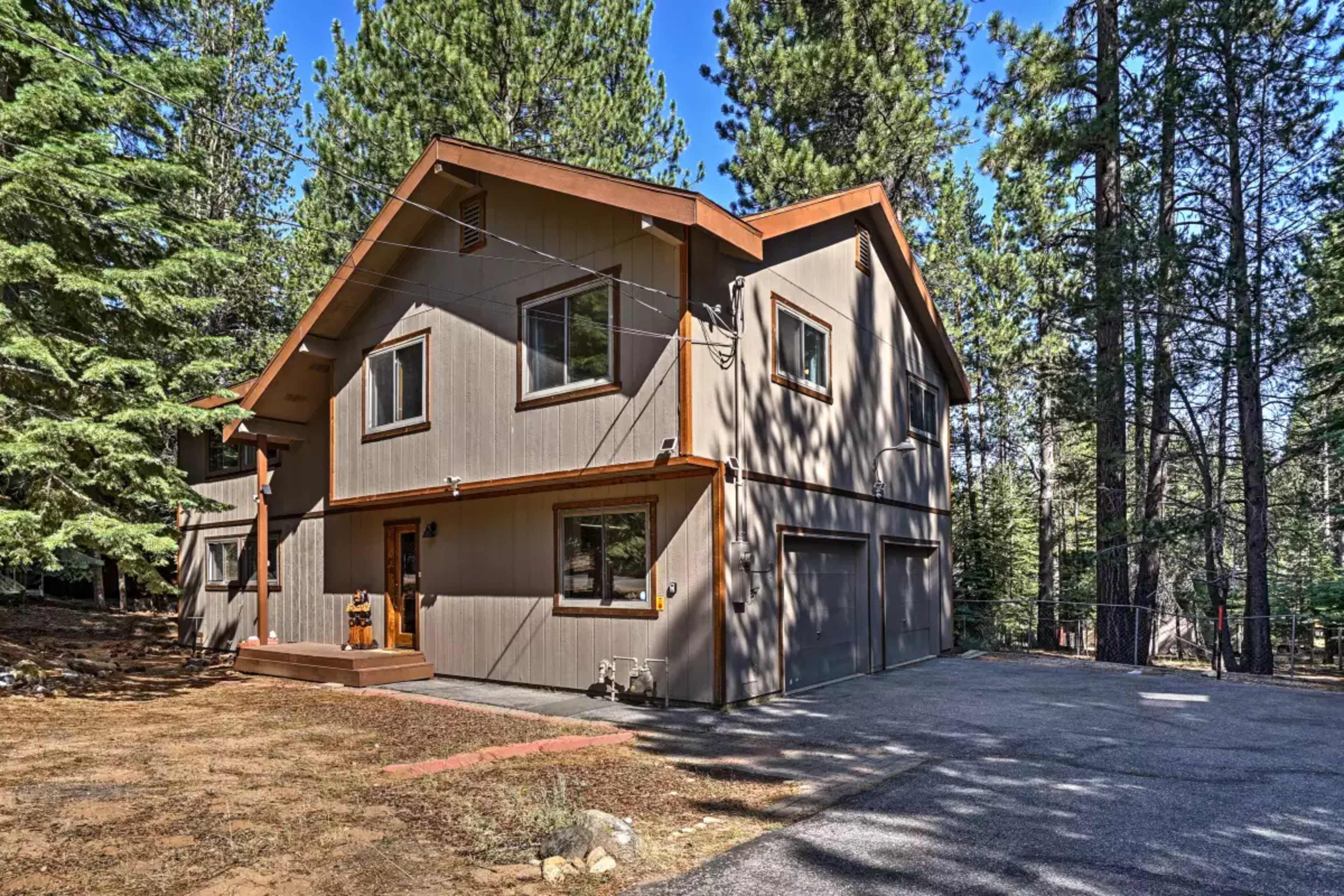 A two-story house with a sloped roof and a wooden front porch is surrounded by tall trees and a gravel driveway.