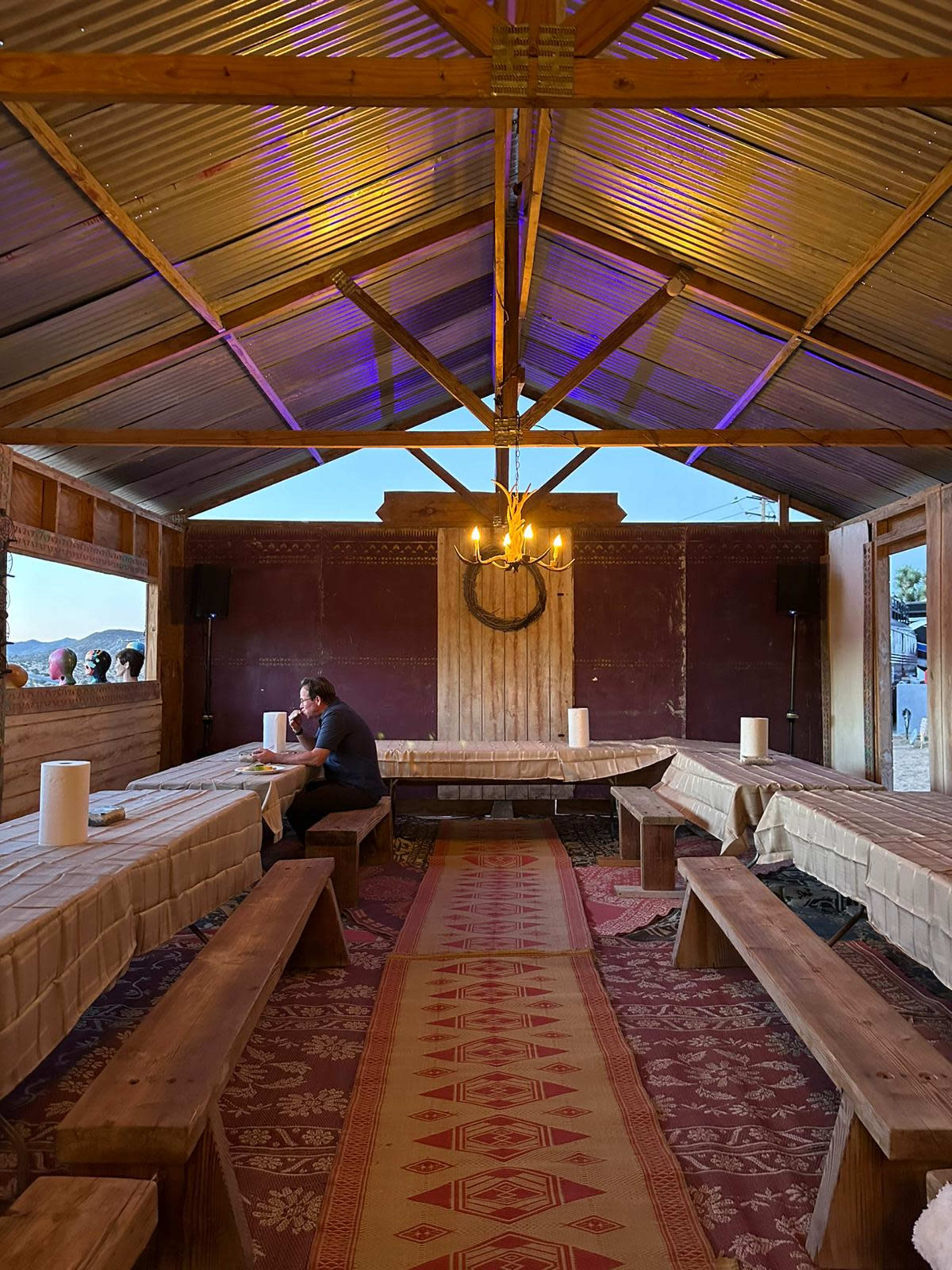 The interior of a rustic venue features long wooden tables and benches, with a central rug and a chandelier, illuminated by soft lighting.