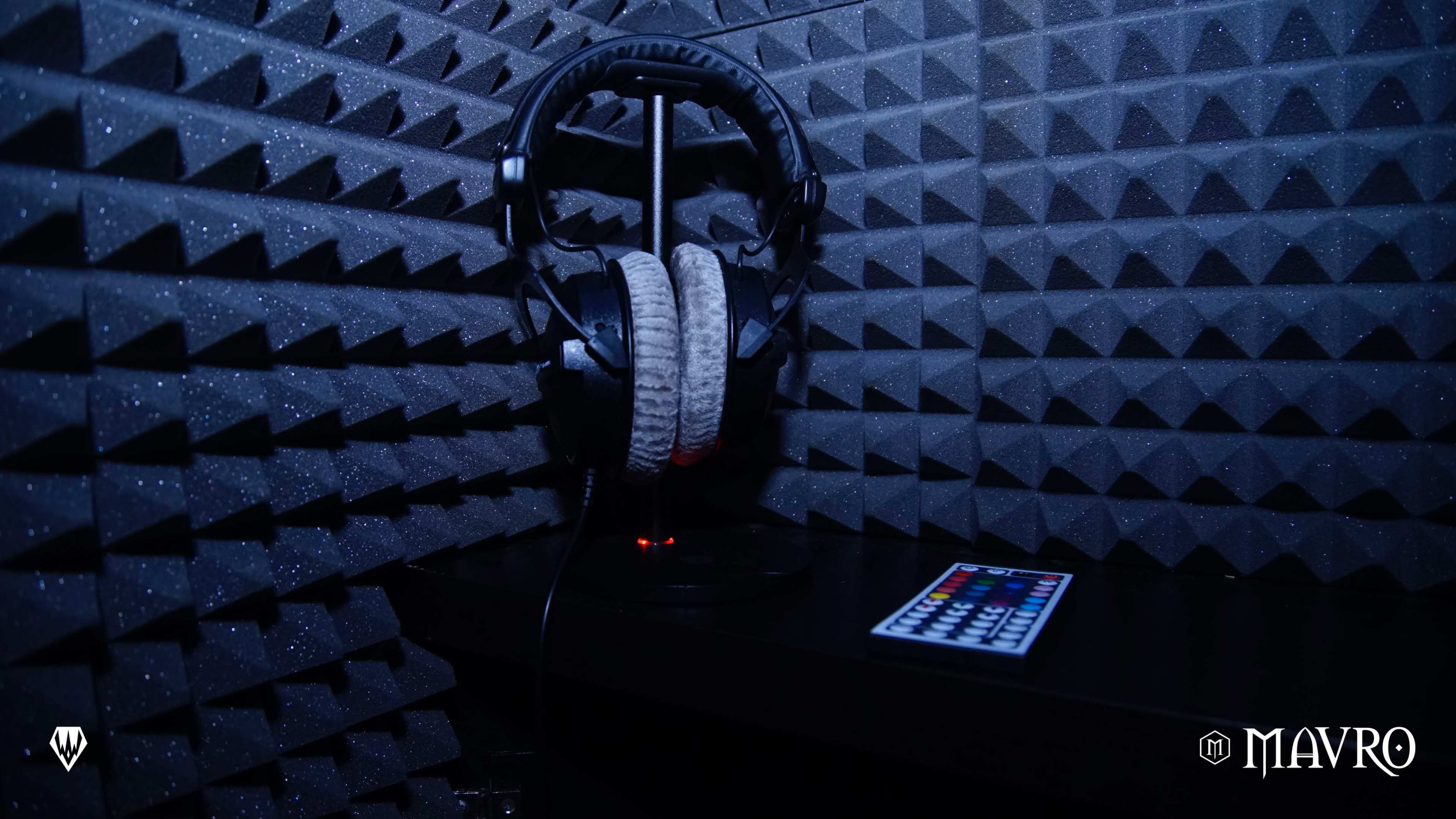 A pair of headphones rests on a stand in a soundproof room, with a remote control placed on a nearby table.