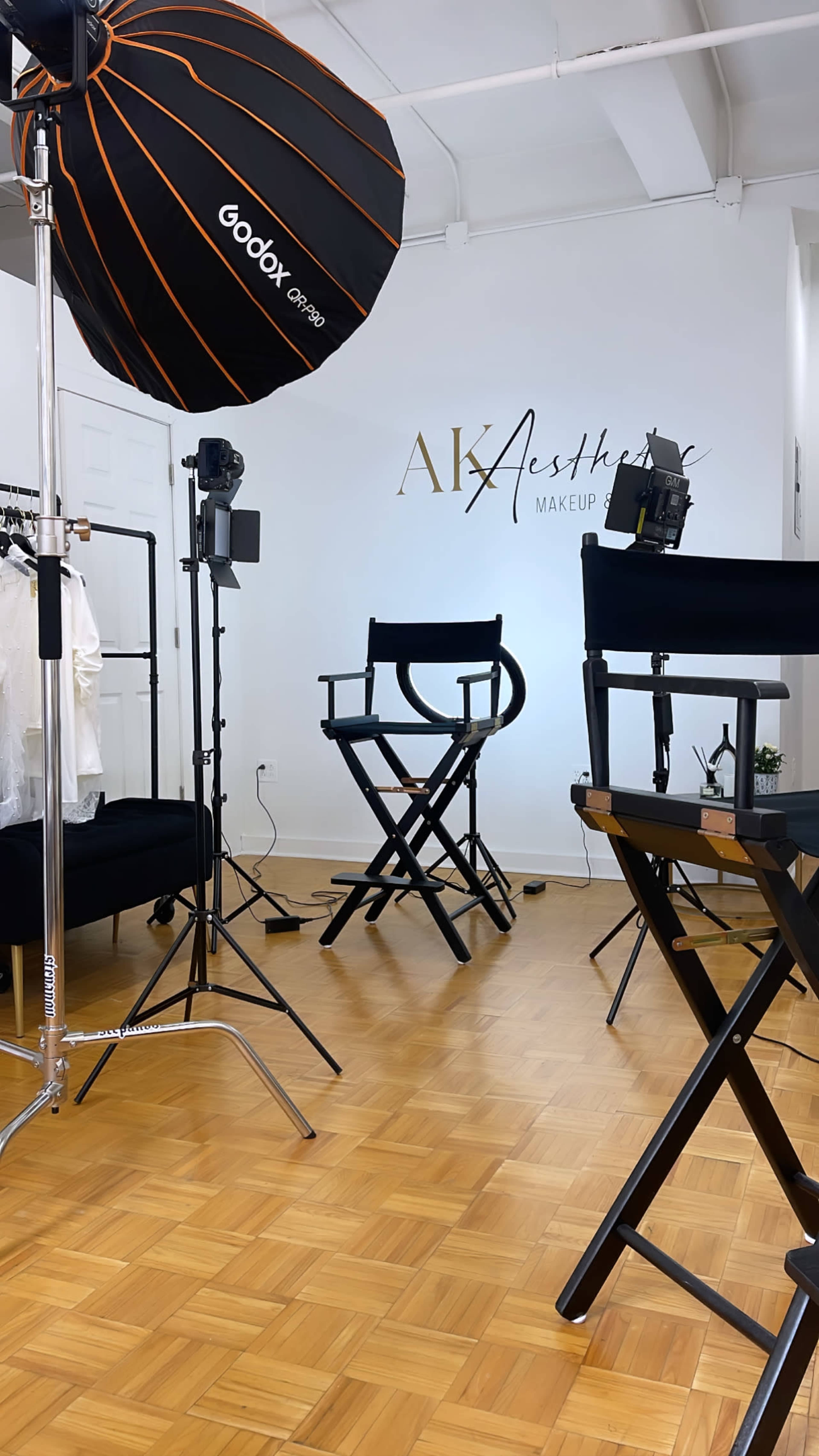 A makeup studio with a large lighting umbrella, director's chairs, and a clothing rack against a wall featuring the name "AK Aesthetics Makeup."
