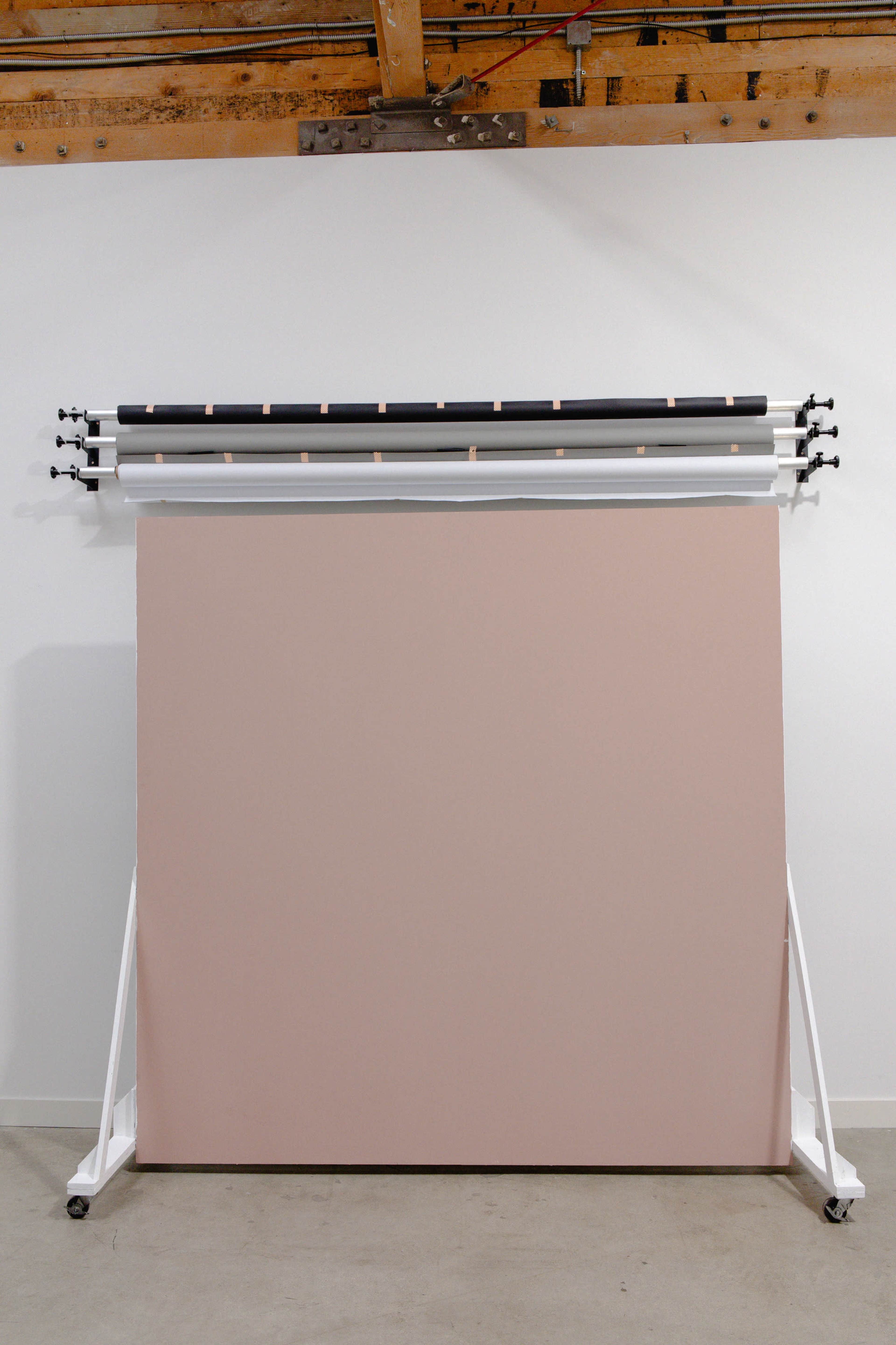 The image shows a simple studio setup featuring a large pink backdrop supported by a white frame, with three rolls of backdrop material hanging above it.