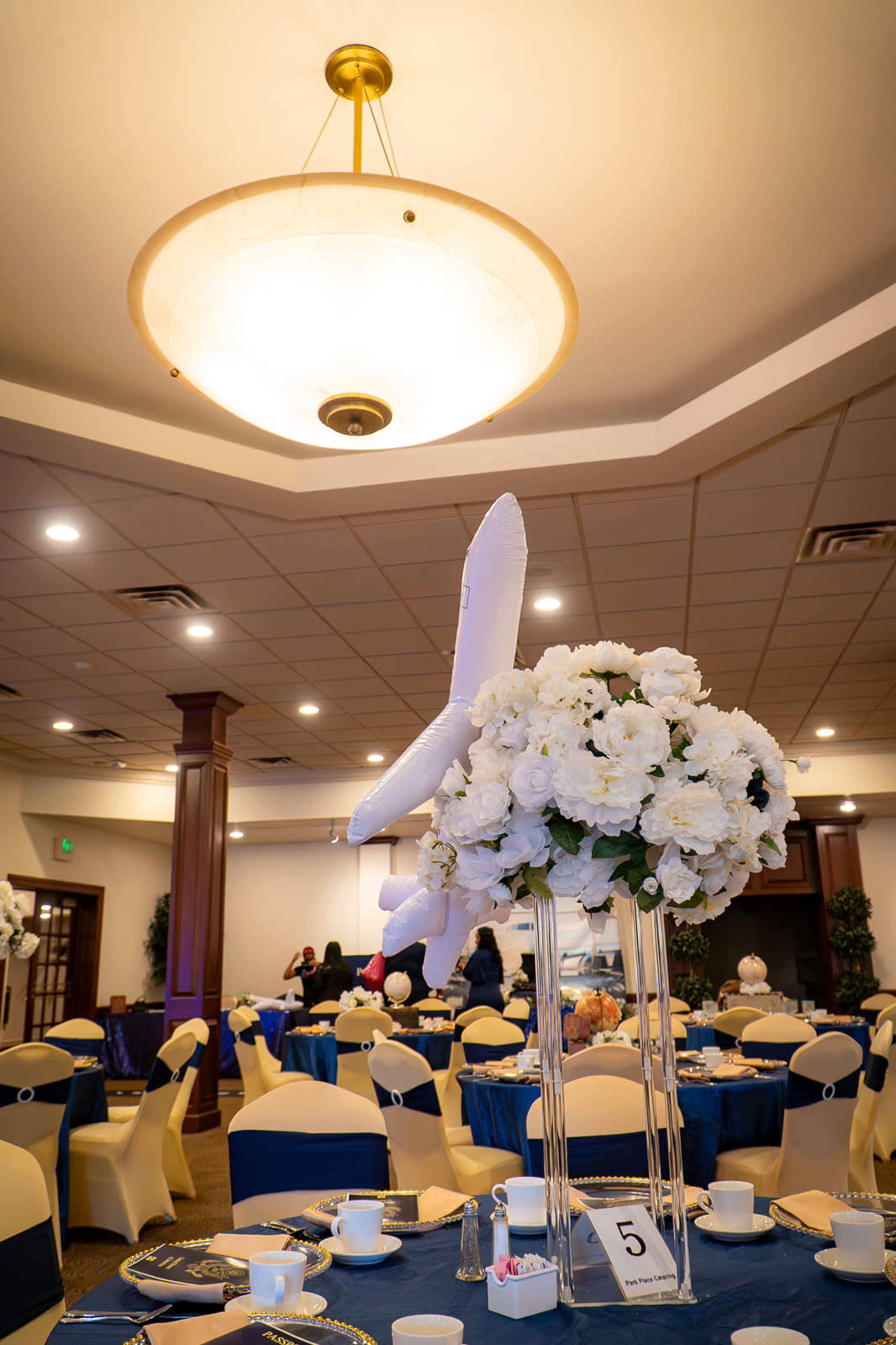 A large floral arrangement in a glass vase sits on a table, surrounded by blue tablecloths and decorated chairs in a banquet hall setting.