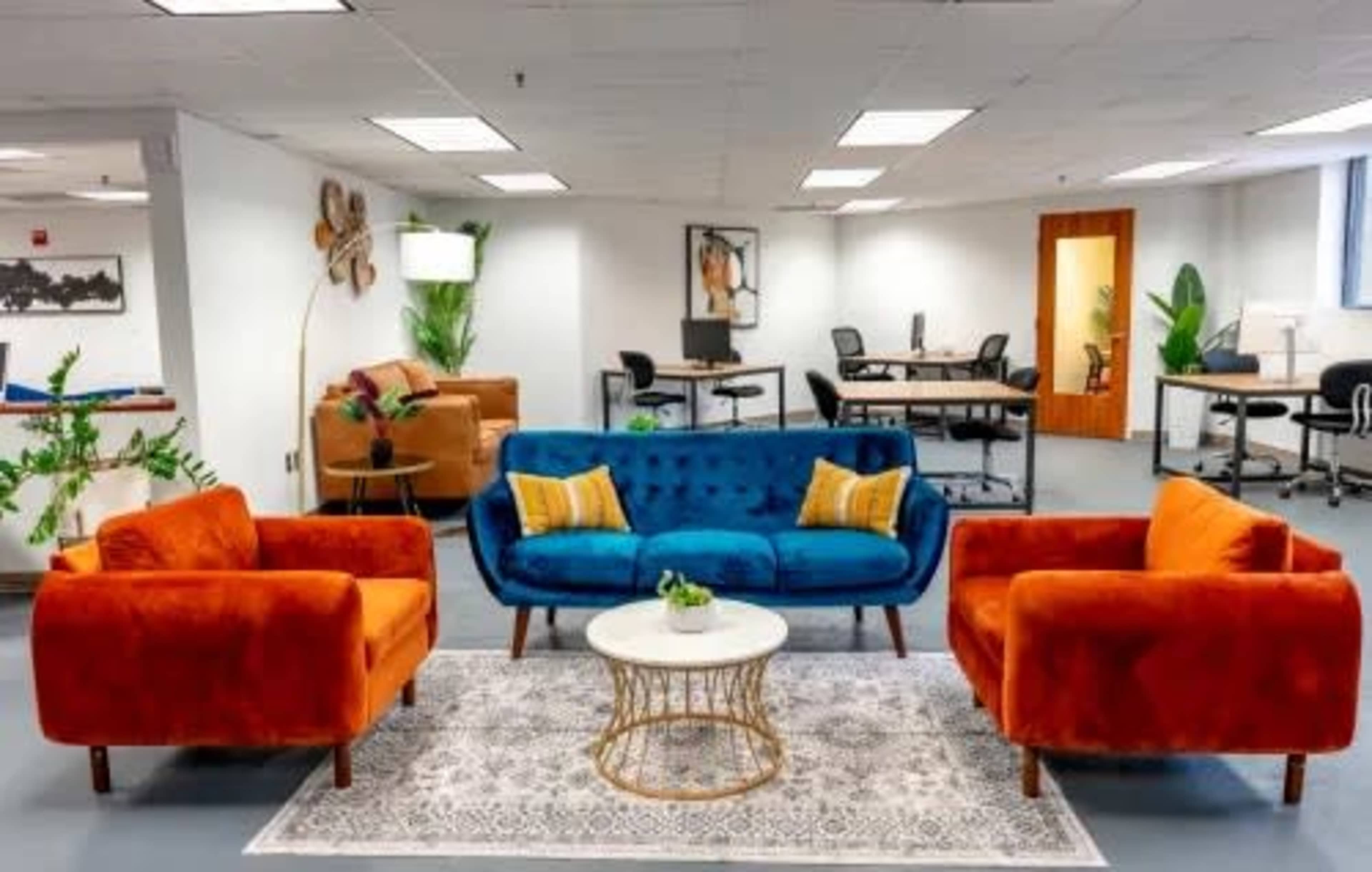 The image shows a modern office lounge area featuring a blue sofa flanked by orange armchairs, a round coffee table, and workspace desks in the background.