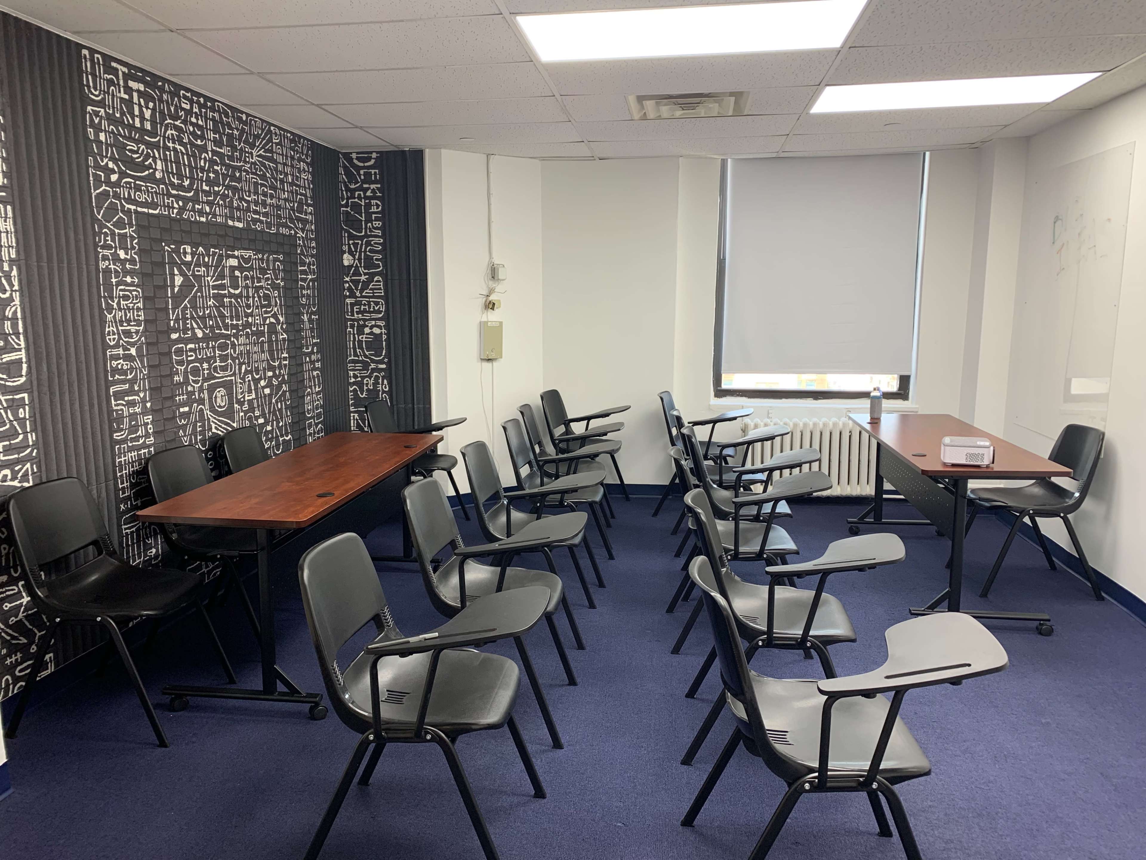 A meeting room with two tables and several black chairs arranged in rows on blue carpeting.