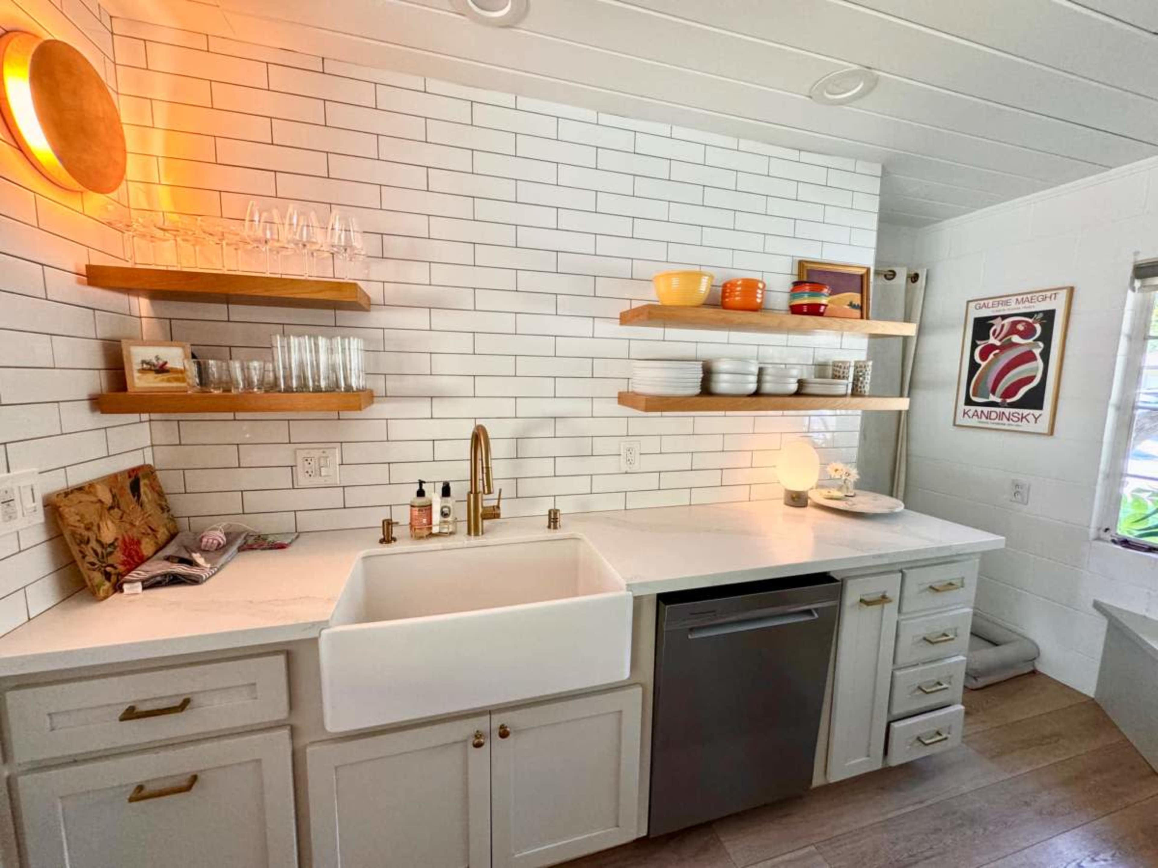 The image shows a modern kitchen with white subway tile walls, wooden shelves holding dishes and glassware, and a farmhouse sink adjacent to a stainless steel dishwasher.