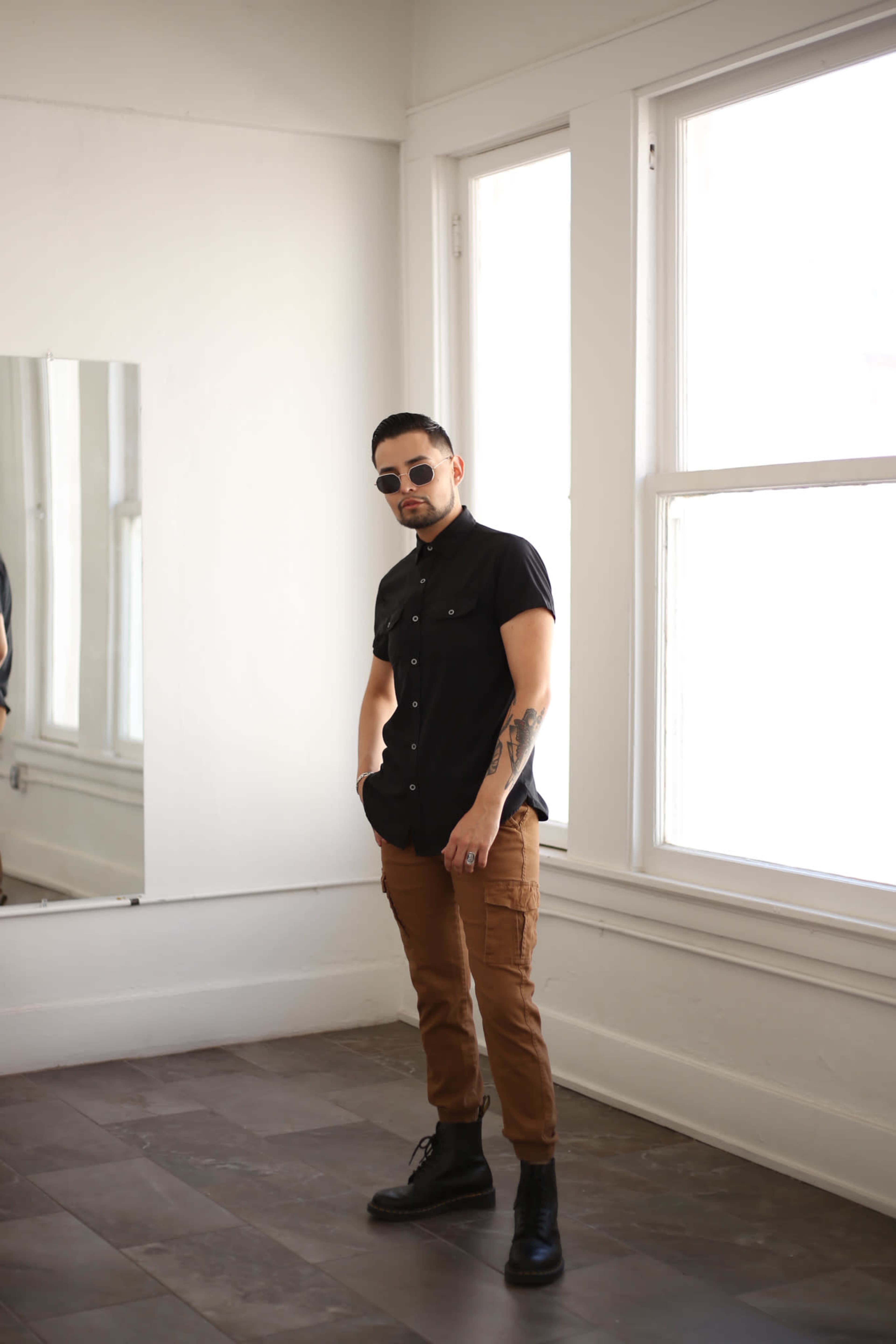 A man wearing sunglasses and a black shirt stands in a room with large windows and a mirror.