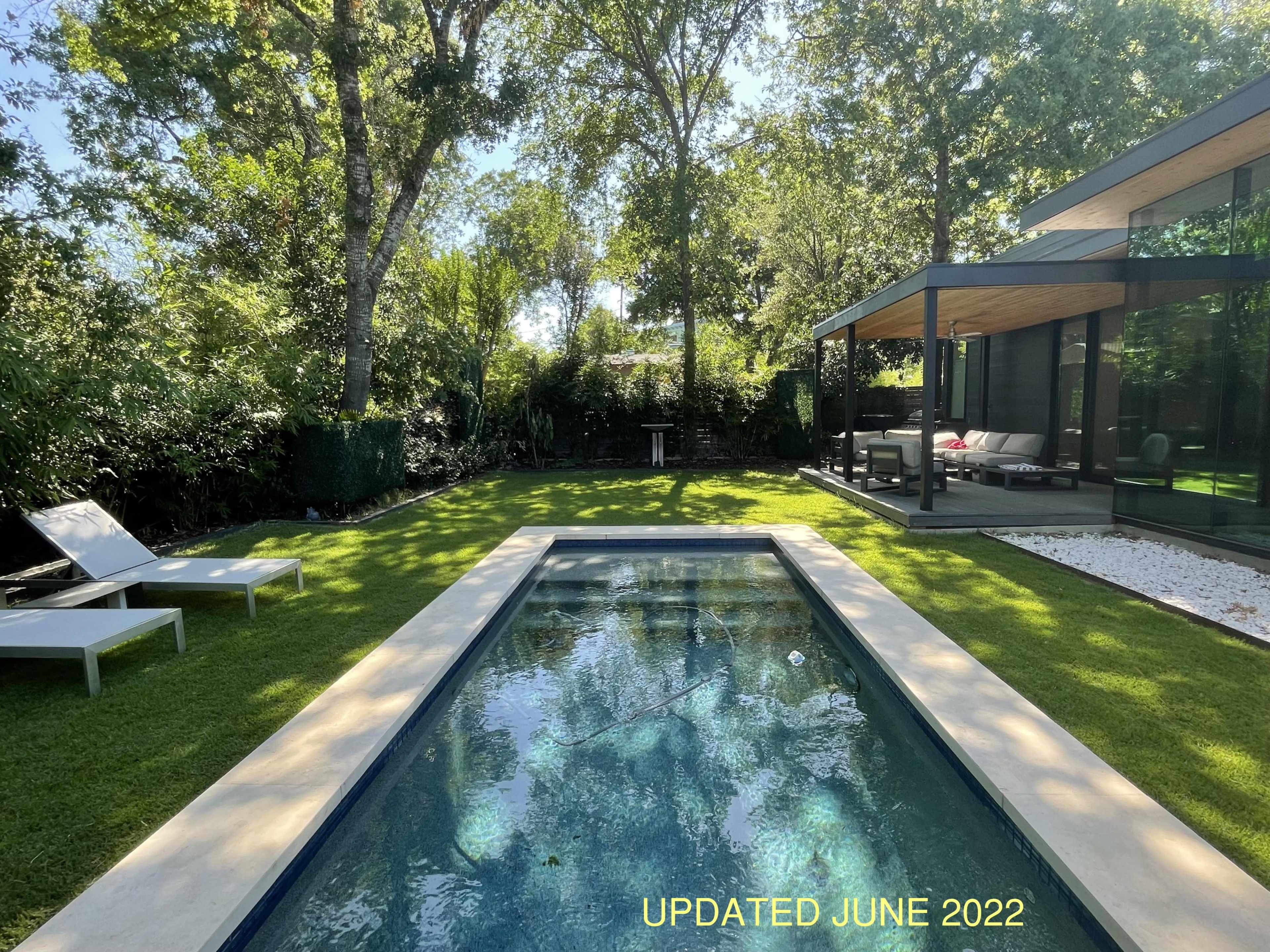 A modern backyard featuring a rectangular pool surrounded by neatly trimmed grass and lounge chairs, alongside a shaded seating area under trees.