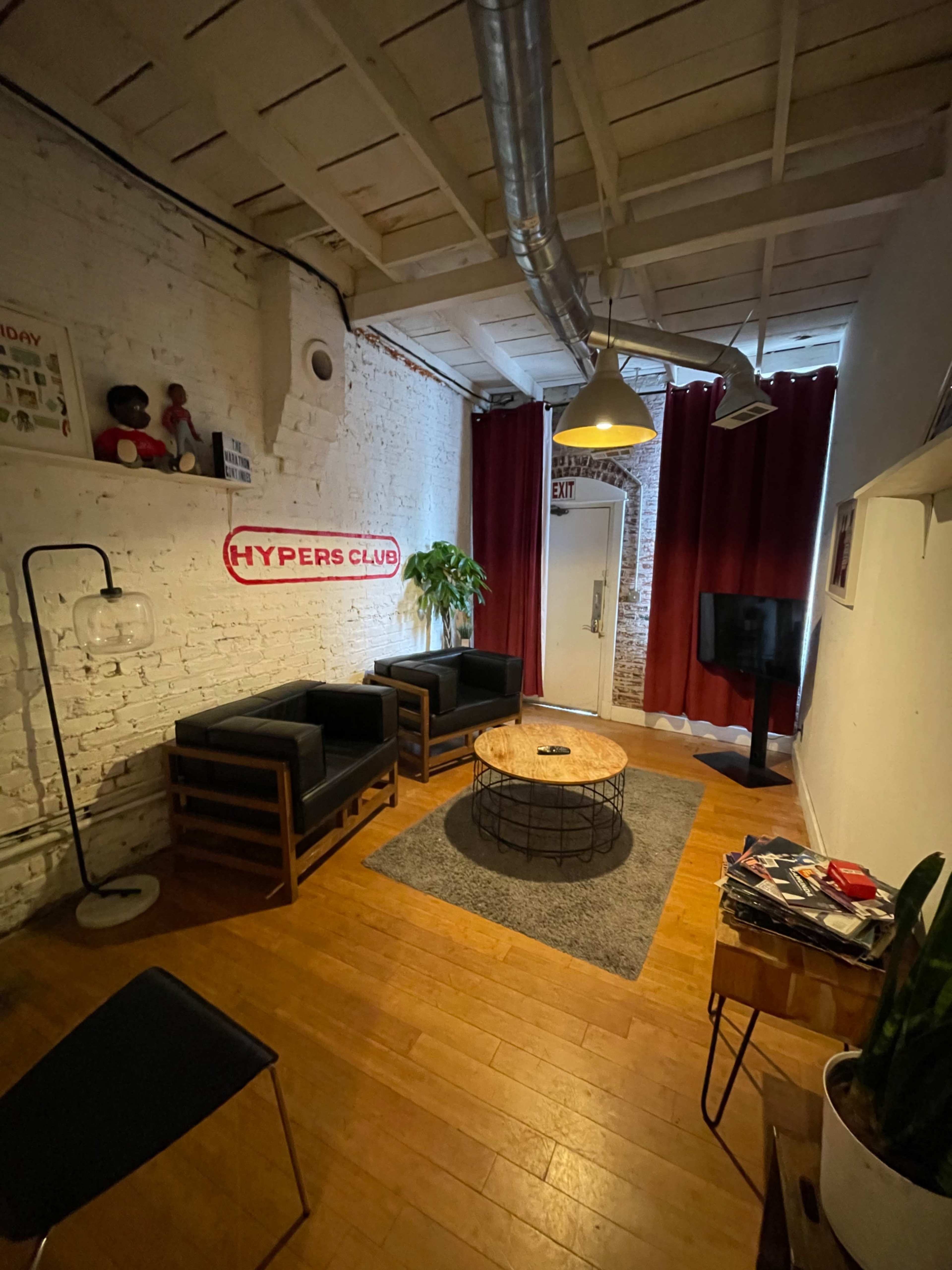 The image shows a cozy lounge area with two black chairs, a round wooden coffee table, a gray rug, and a plant, complemented by red curtains and exposed brick walls.