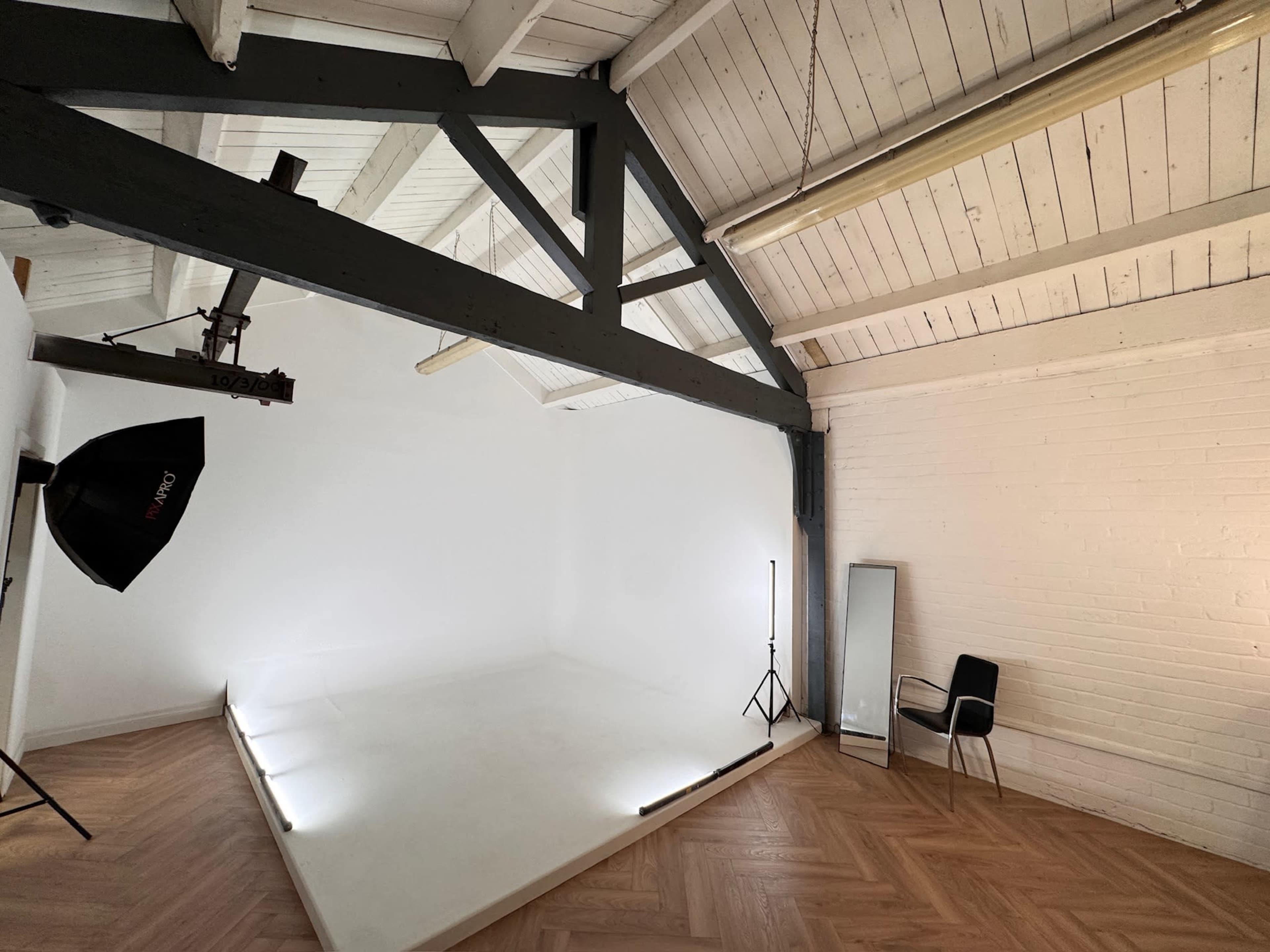 The image shows a minimalist studio space with a white backdrop, wooden ceiling beams, and a chair positioned near a mirror.