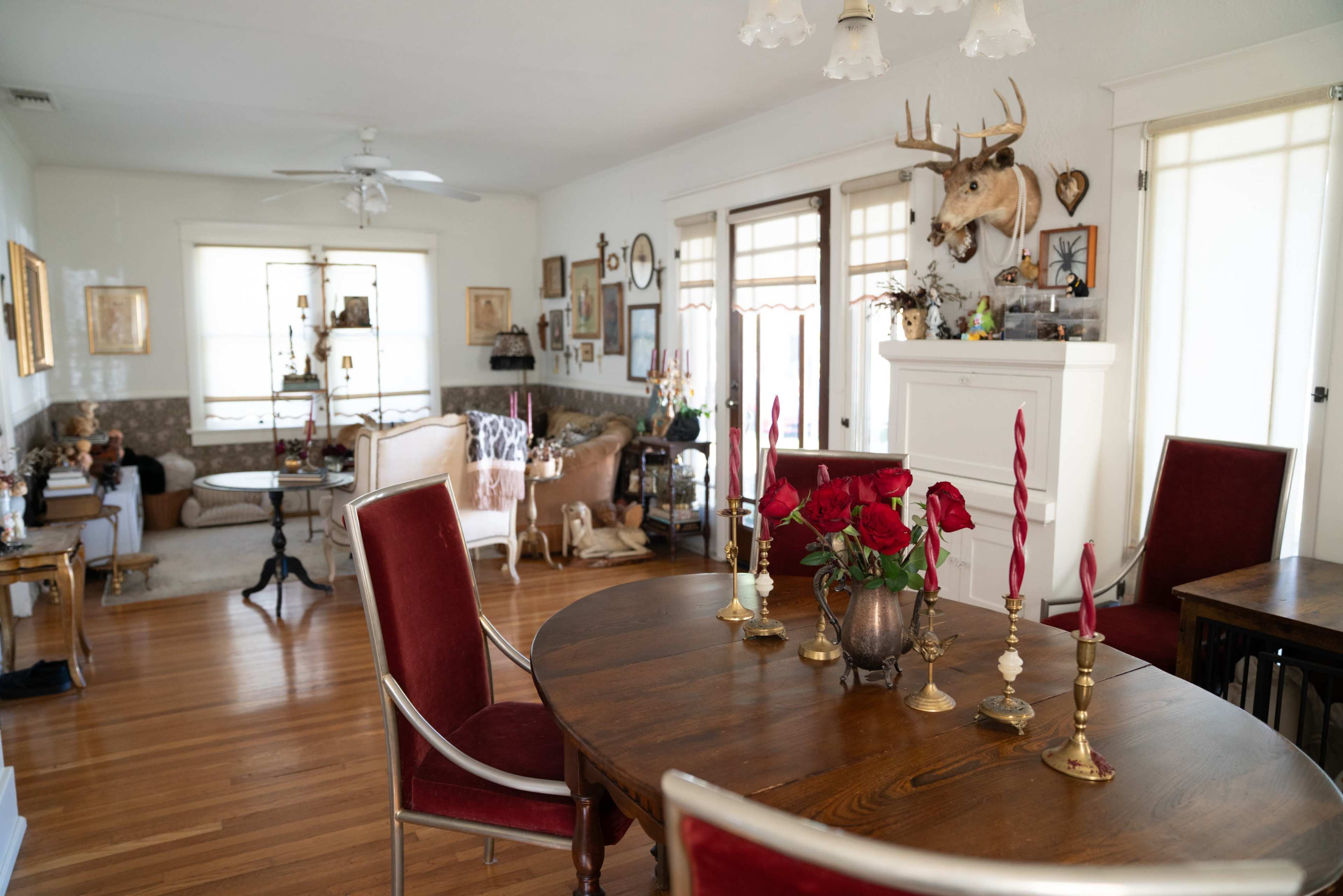 The image shows a cozy dining area with a round wooden table set for a meal, surrounded by red upholstered chairs, while the adjacent living space features various decorative items and a mounted deer head on the wall.