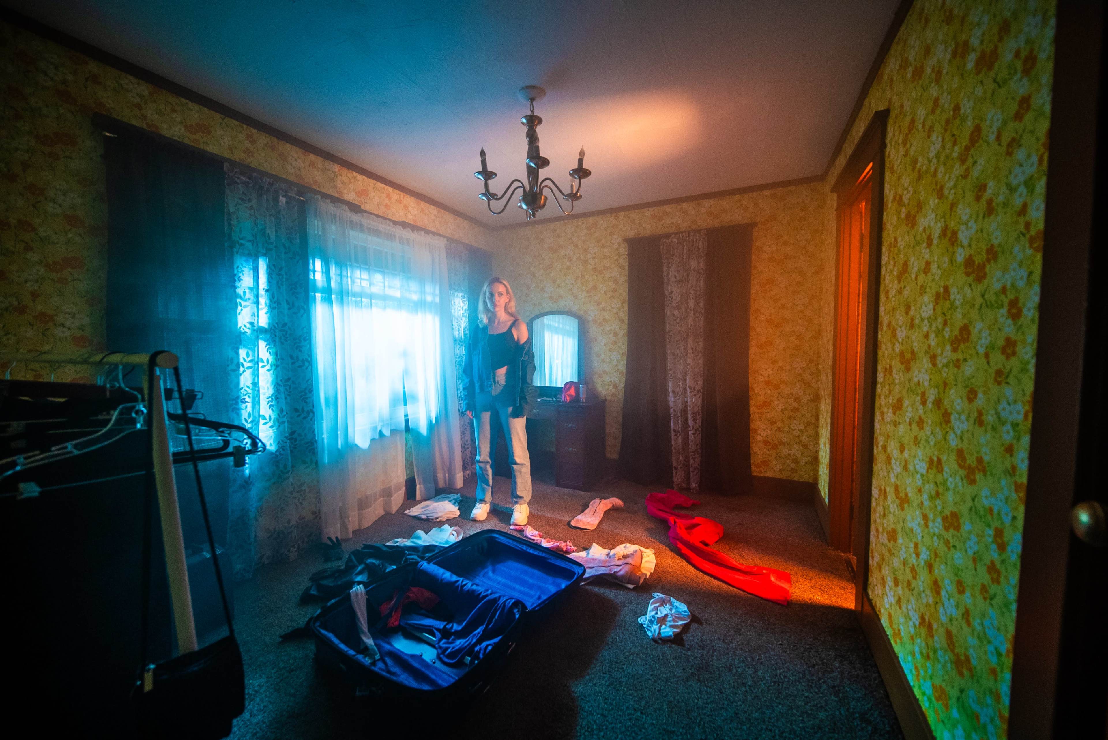 A woman stands in a dimly lit room surrounded by scattered clothes and a partially unpacked suitcase.
