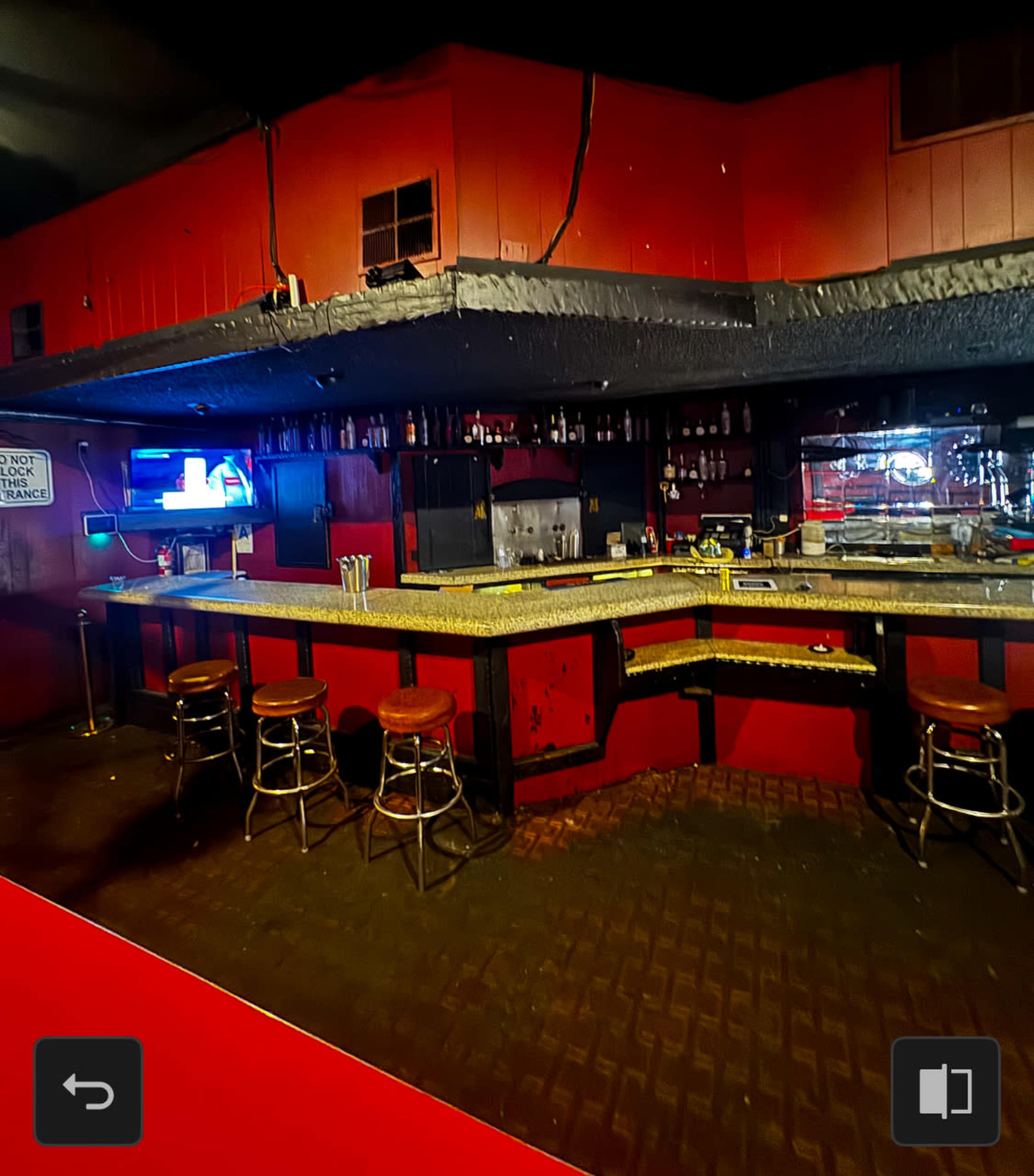 The image shows an empty bar area with a granite countertop, several bar stools, and shelves filled with bottles, set against a backdrop of red walls and dim lighting.