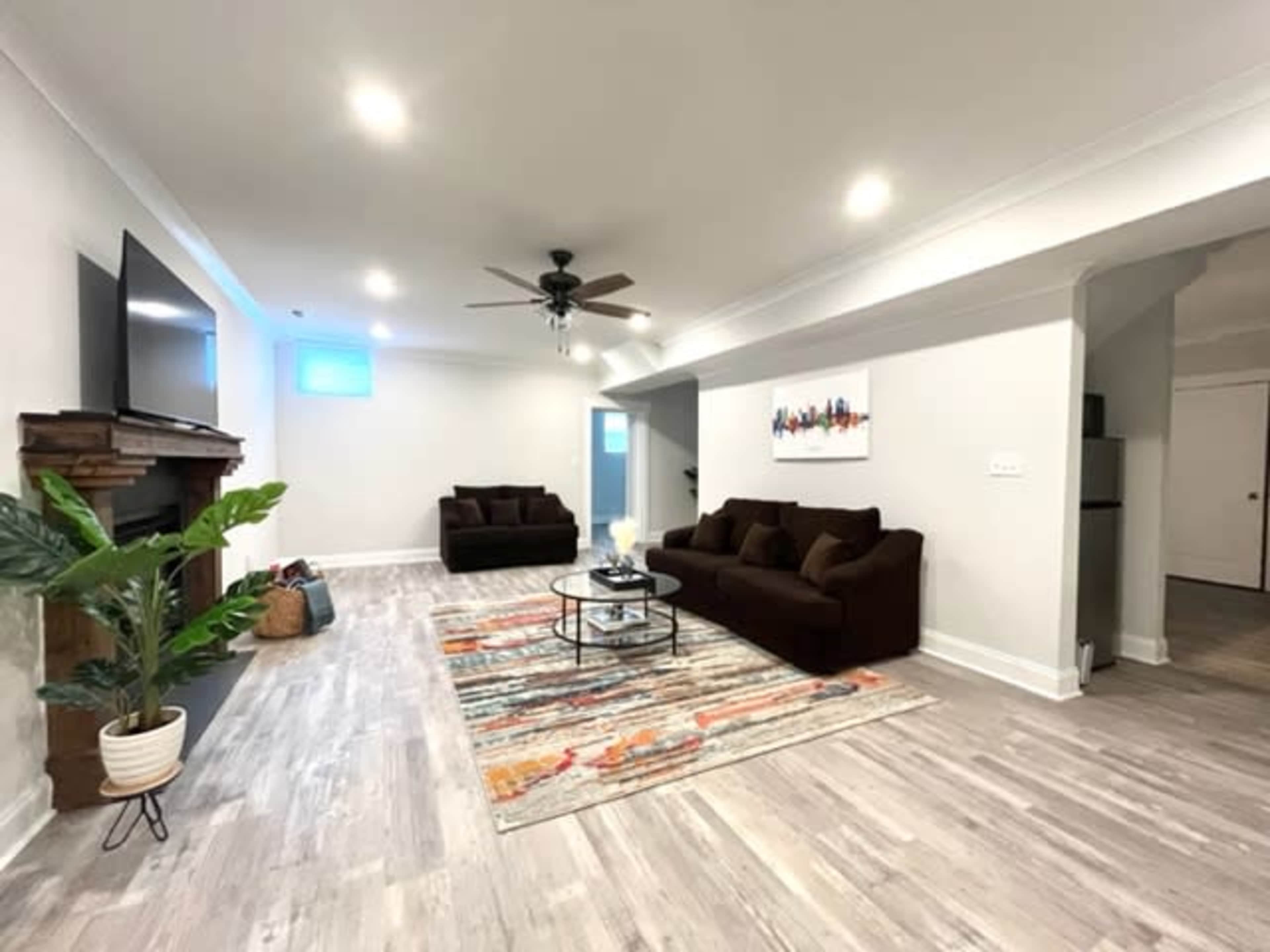 A spacious living room features two dark brown sofas, a coffee table on a colorful rug, and a television mounted on the wall, with a ceiling fan and a plant in the corner.