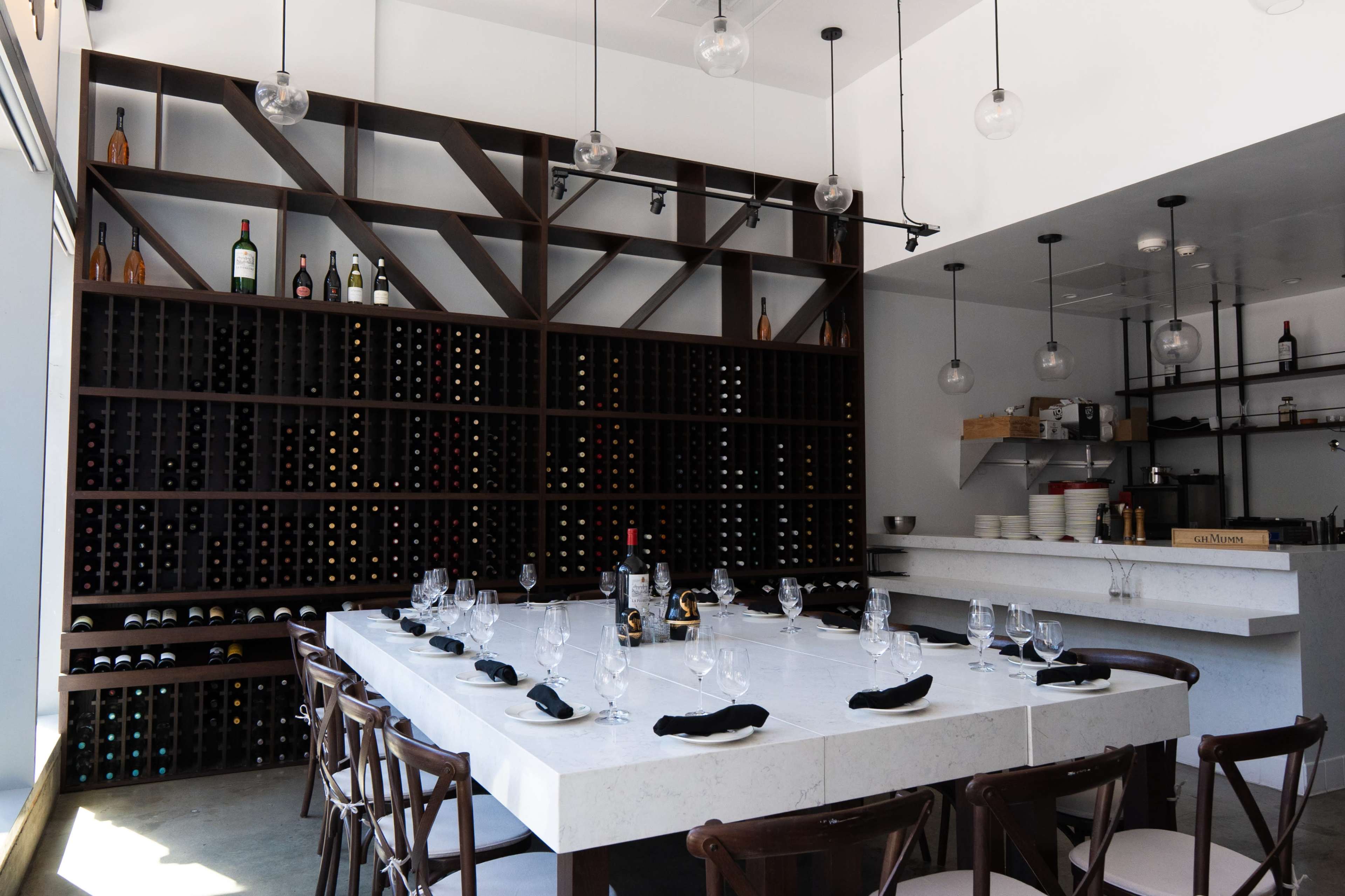 The image shows a modern dining room with a large marble table set for a meal, surrounded by wooden chairs, and a wall of wine racks filled with various bottles.