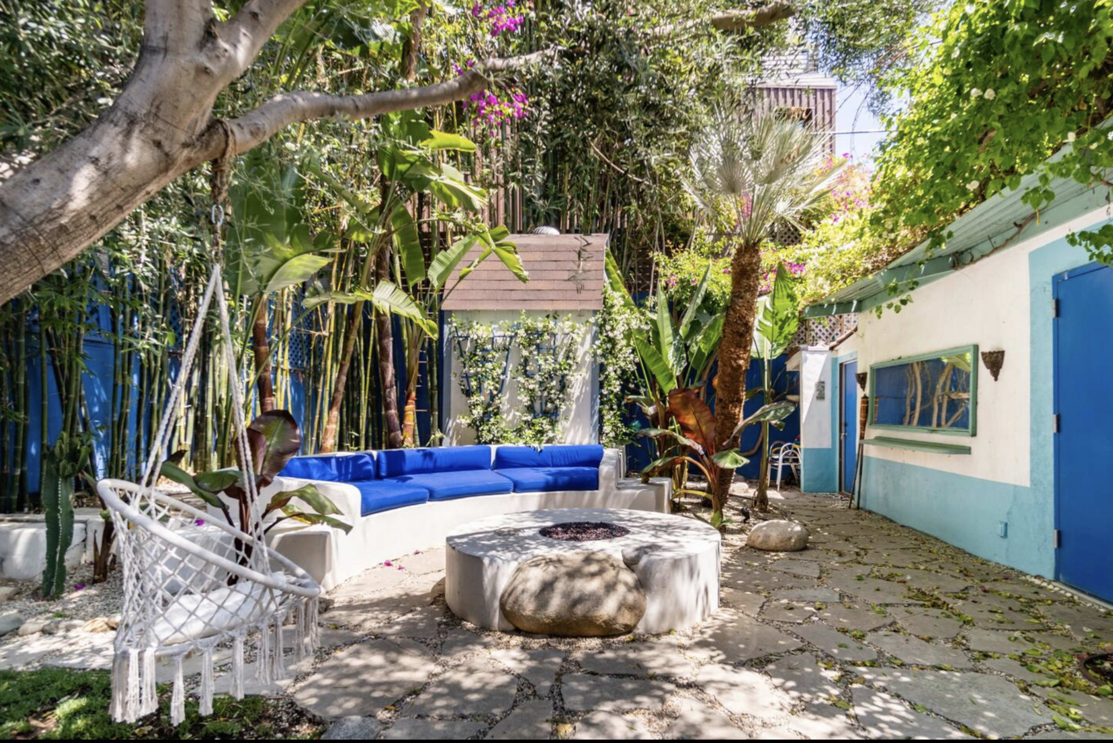 A tranquil outdoor space features a circular stone fire pit surrounded by a curved bench and hanging swings, with lush greenery and colorful flowers in the background.