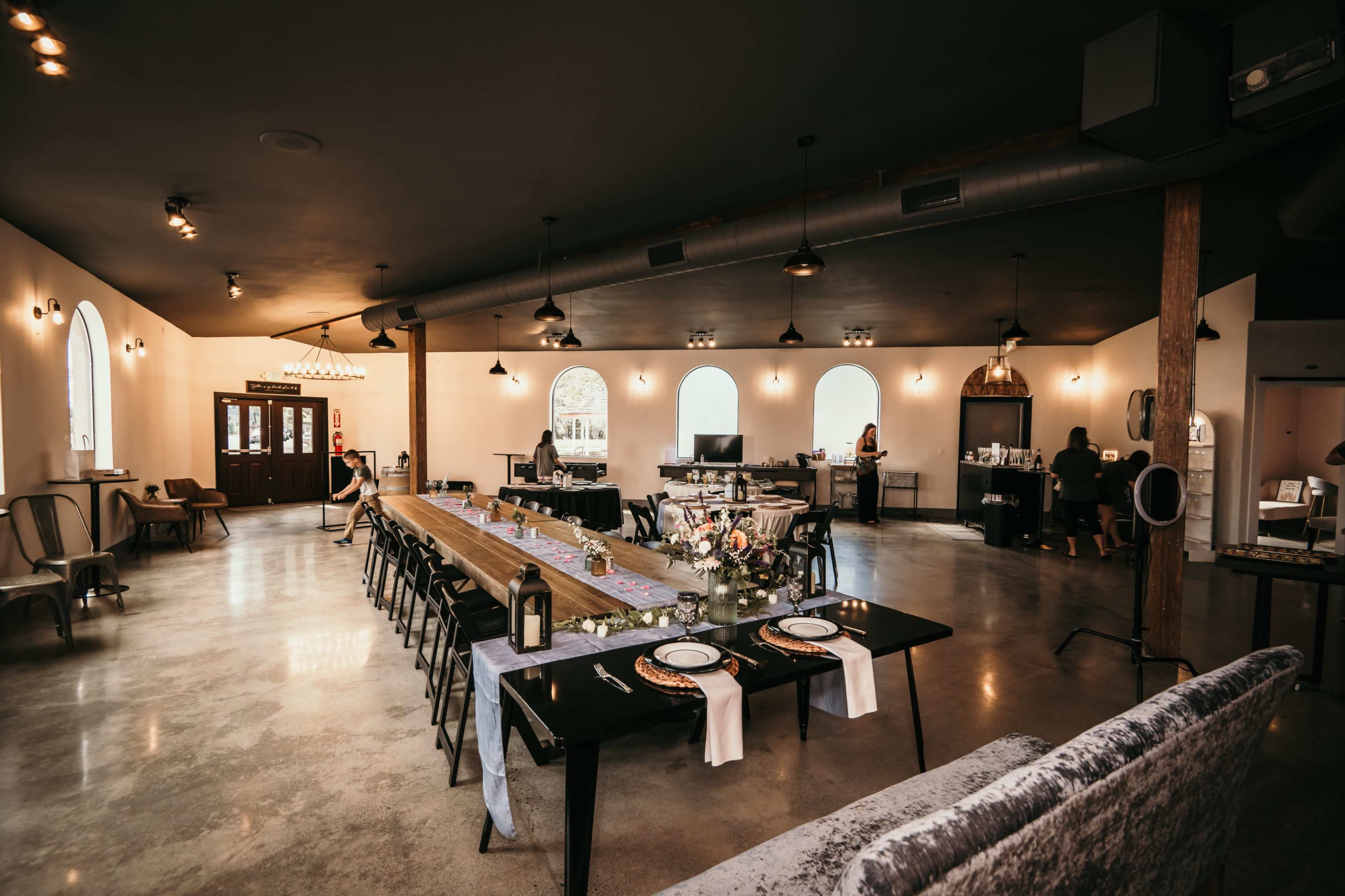 A spacious dining area features a long wooden table set with plates and floral arrangements, surrounded by chairs, with large windows letting in natural light.