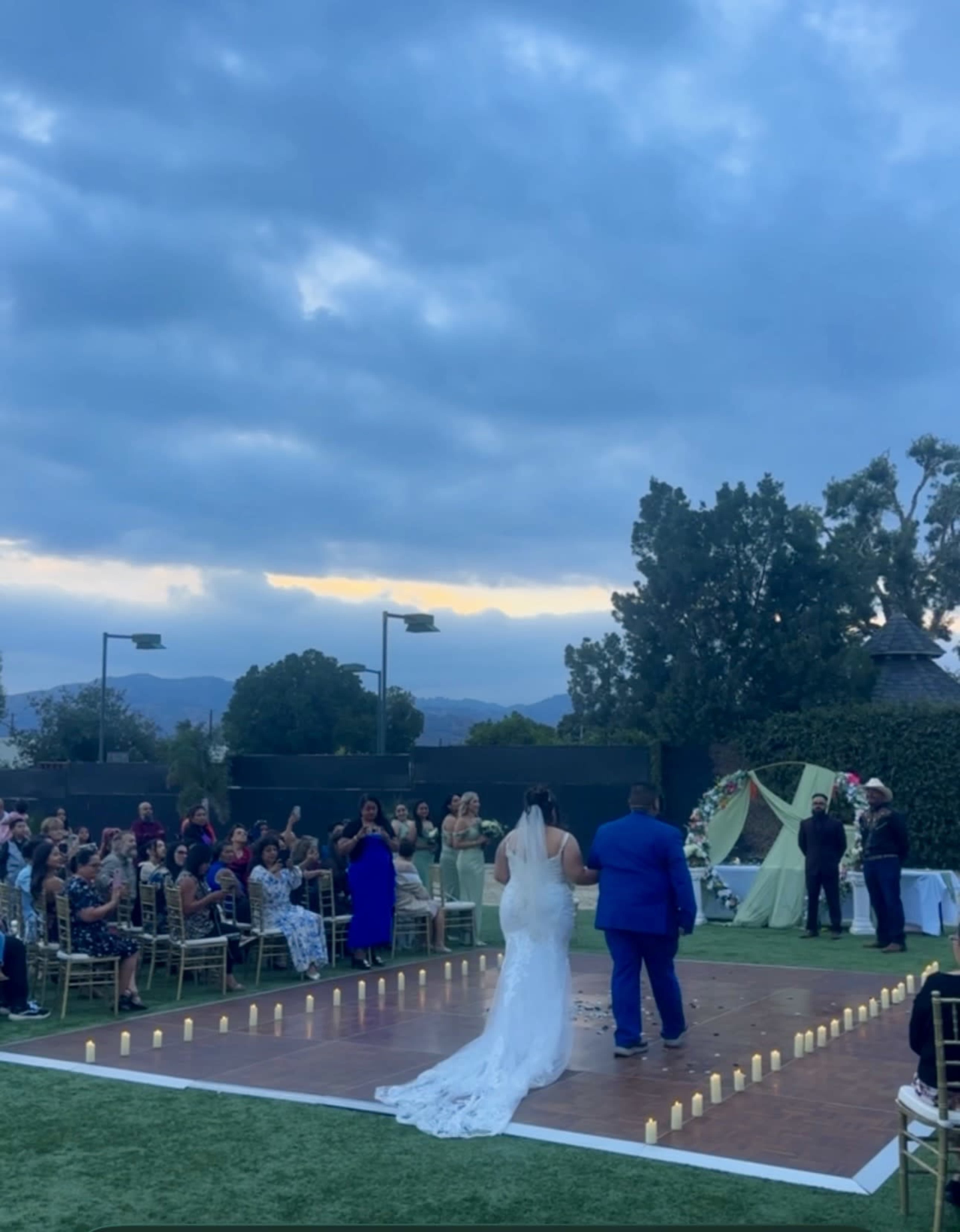 A bride and groom stand on a decorative platform during an outdoor wedding ceremony, surrounded by seated guests and candlelit pathways.