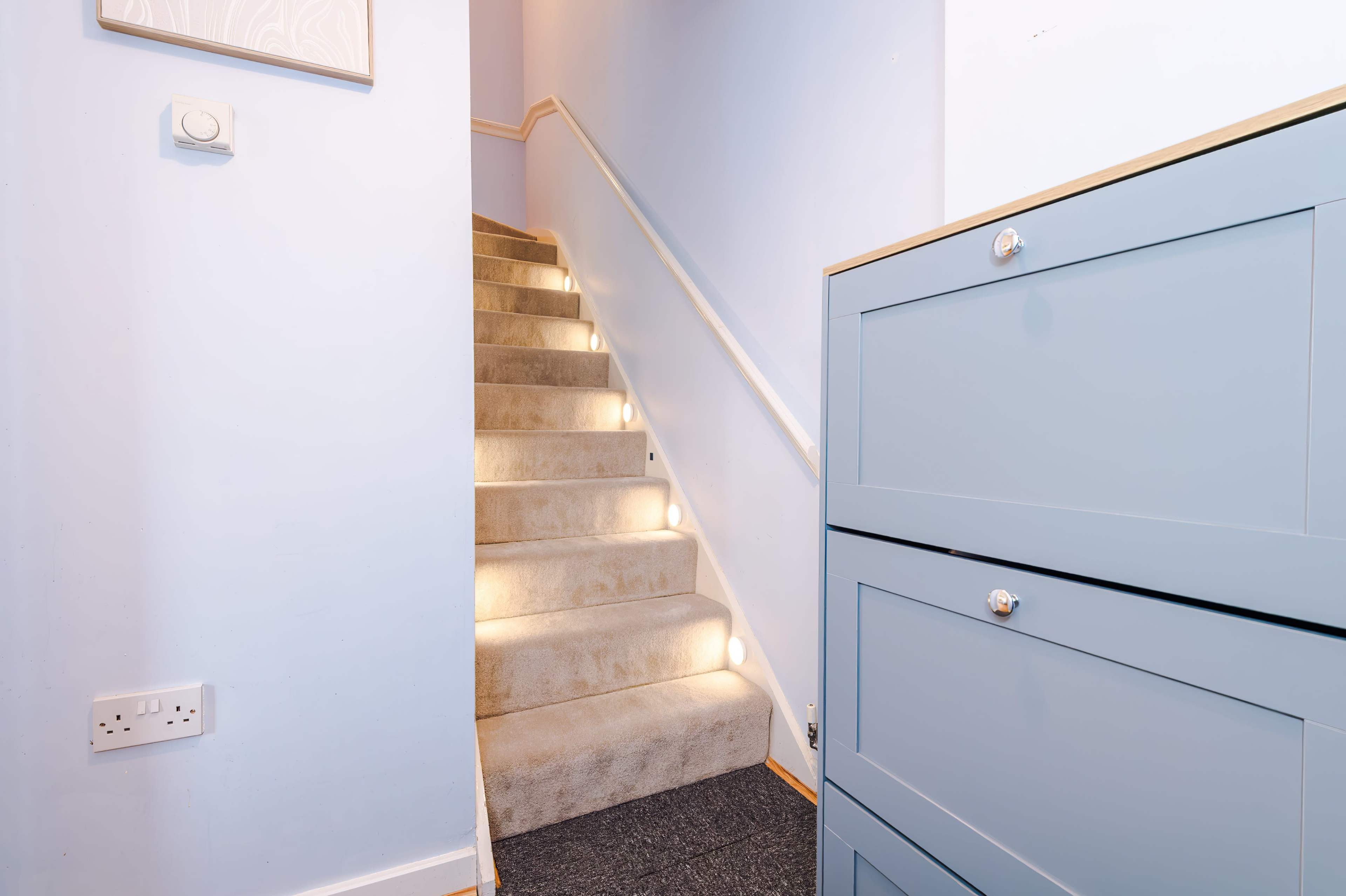 The image shows a staircase with lighted steps, leading up, next to a light blue storage cabinet against a wall.