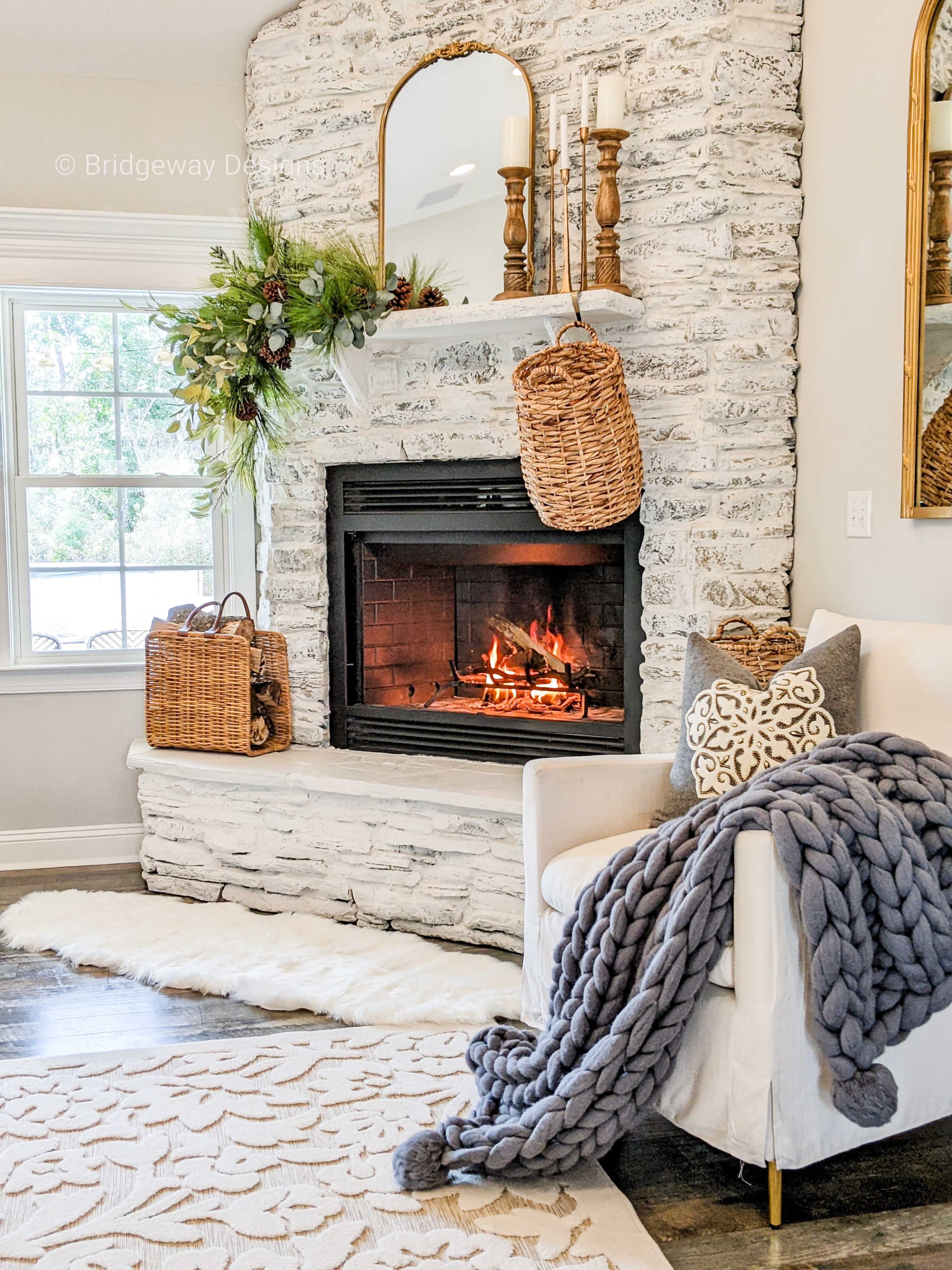 A cozy living room features a stone fireplace with a warm fire, surrounded by decorative elements like a mirror, candles, and woven baskets.