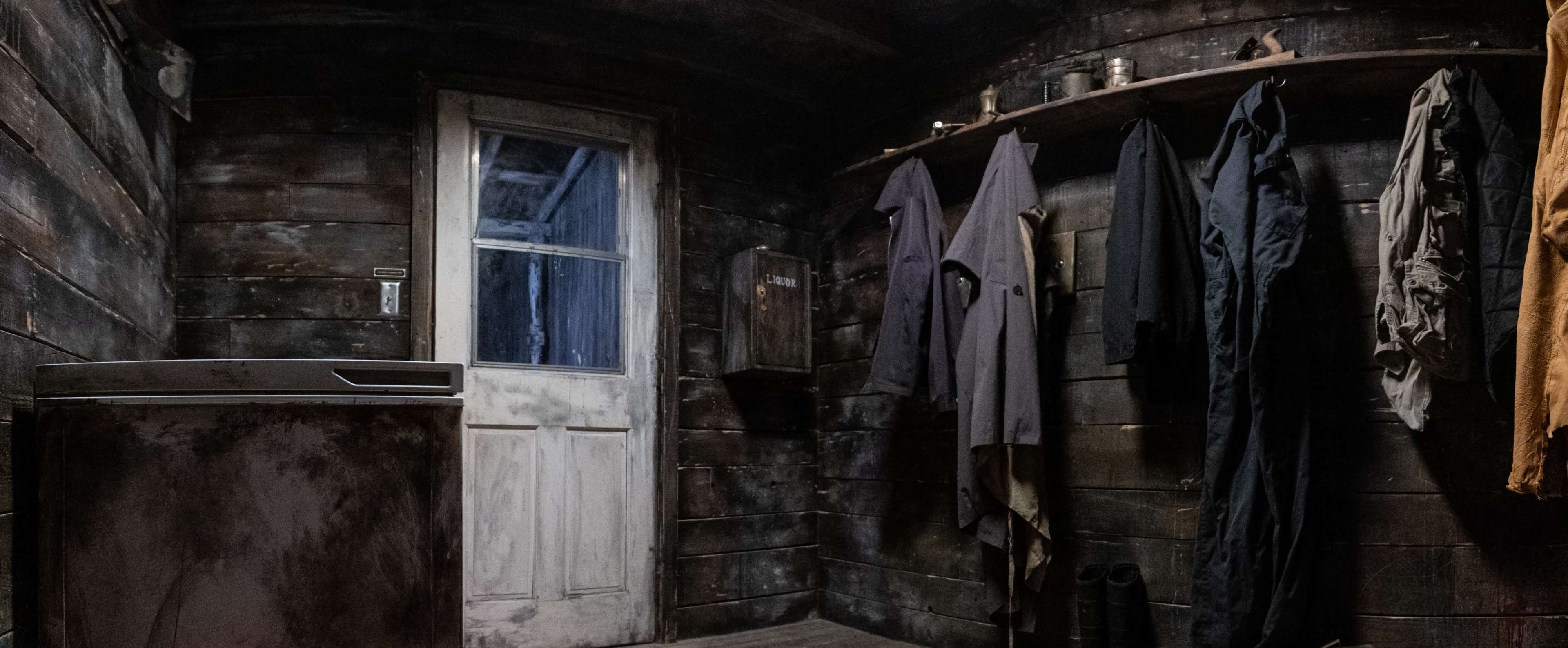 A dimly lit room with wooden walls, featuring a refrigerator, a door, and several garments hanging on a shelf.
