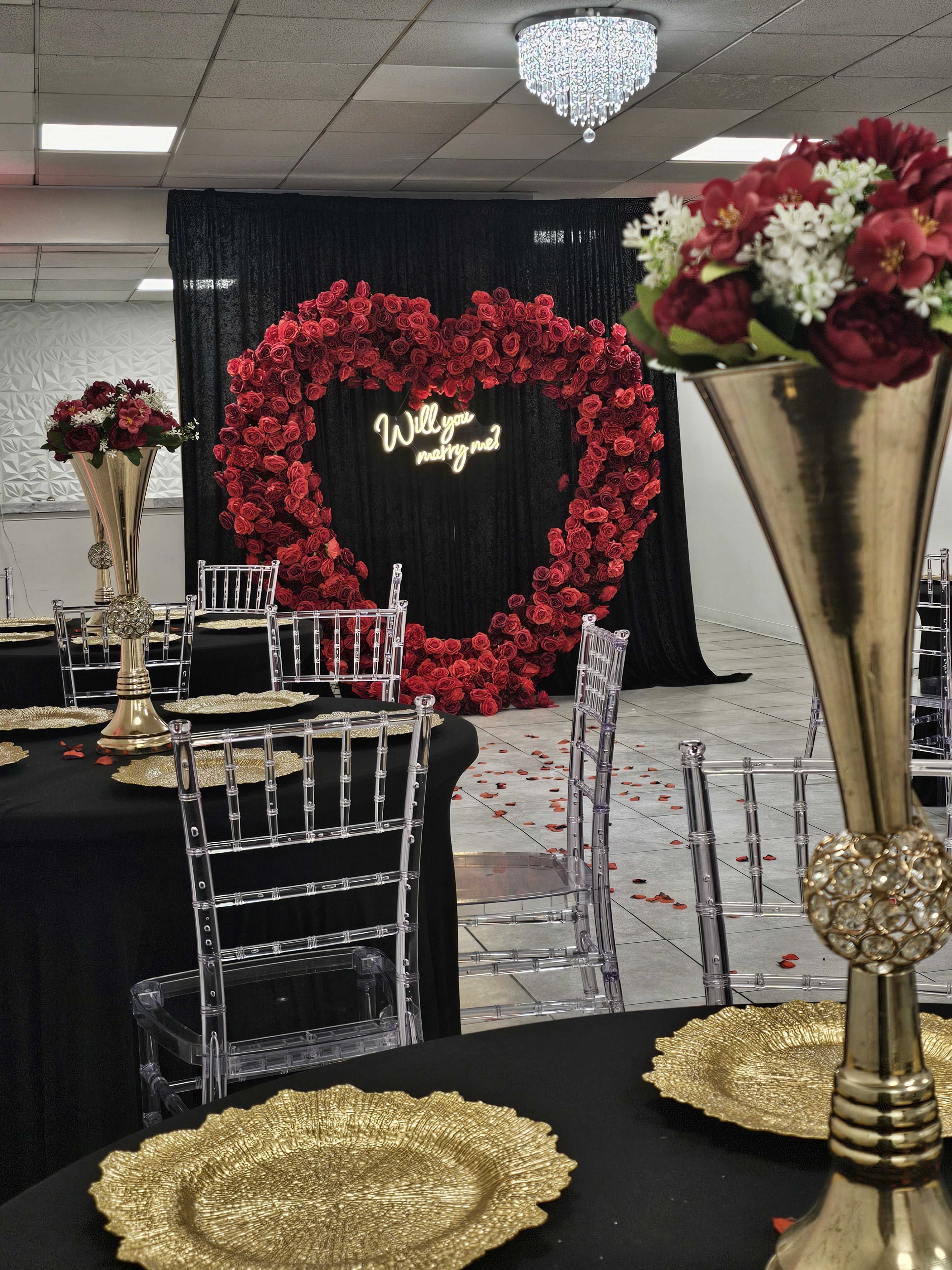 The image shows a decorated event space featuring tables with gold accents, clear chairs, and a heart-shaped backdrop made of red flowers with the phrase "Will you marry me?" on it.