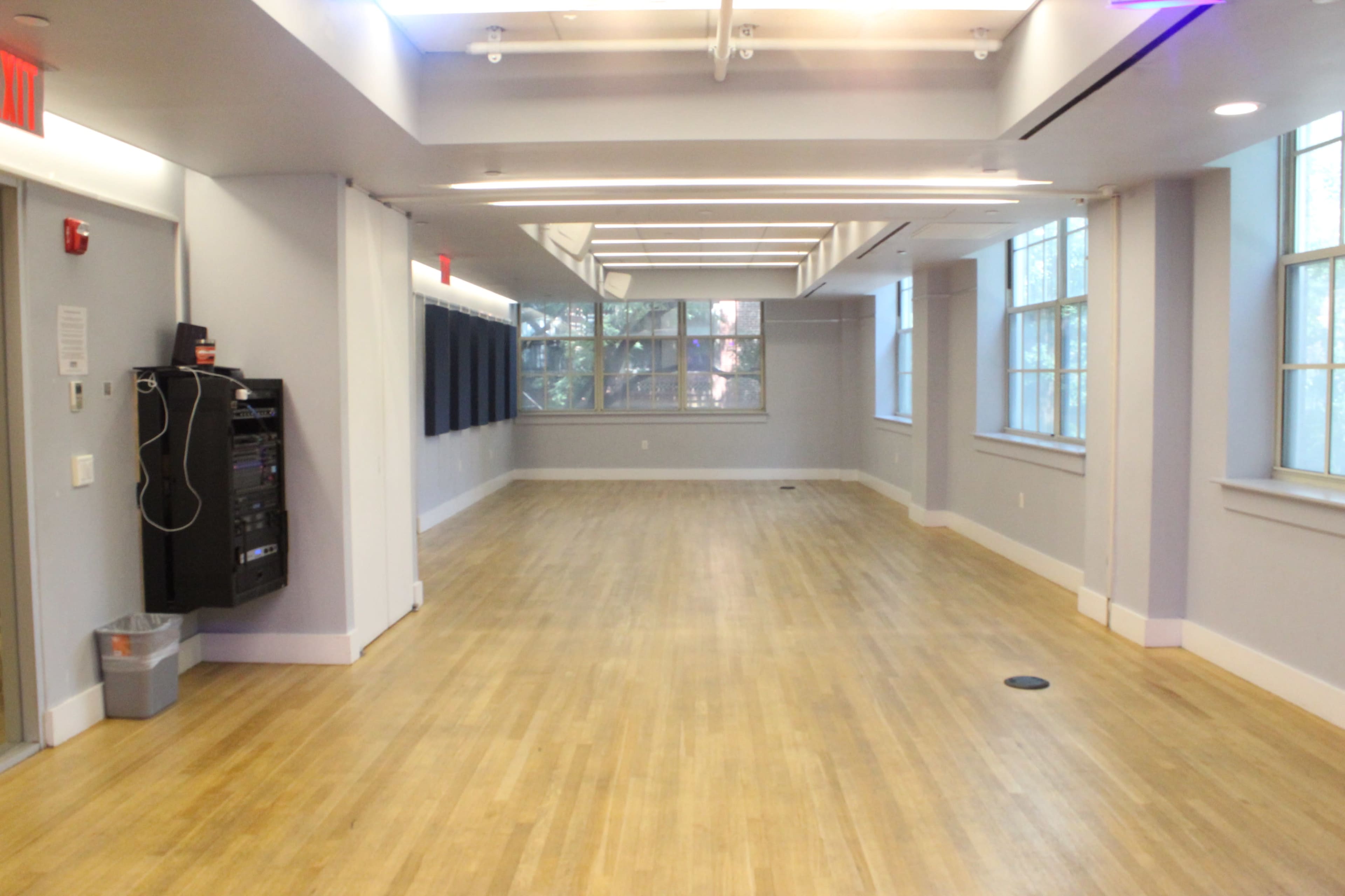 The image shows a spacious, well-lit interior room with wooden flooring and large windows lining one wall.