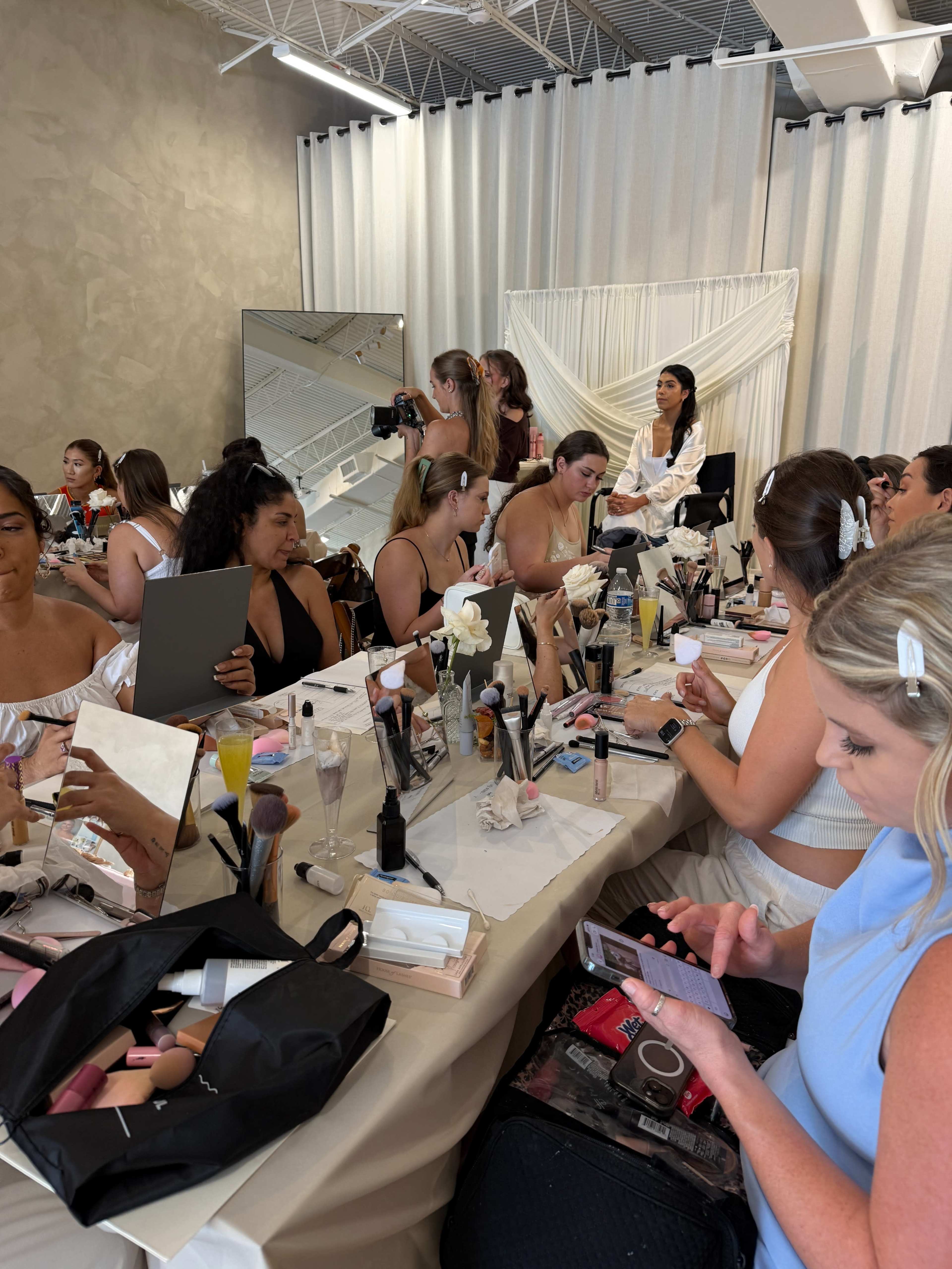 A group of people are seated at tables in a beauty studio, applying makeup while some take photos and others use their phones.