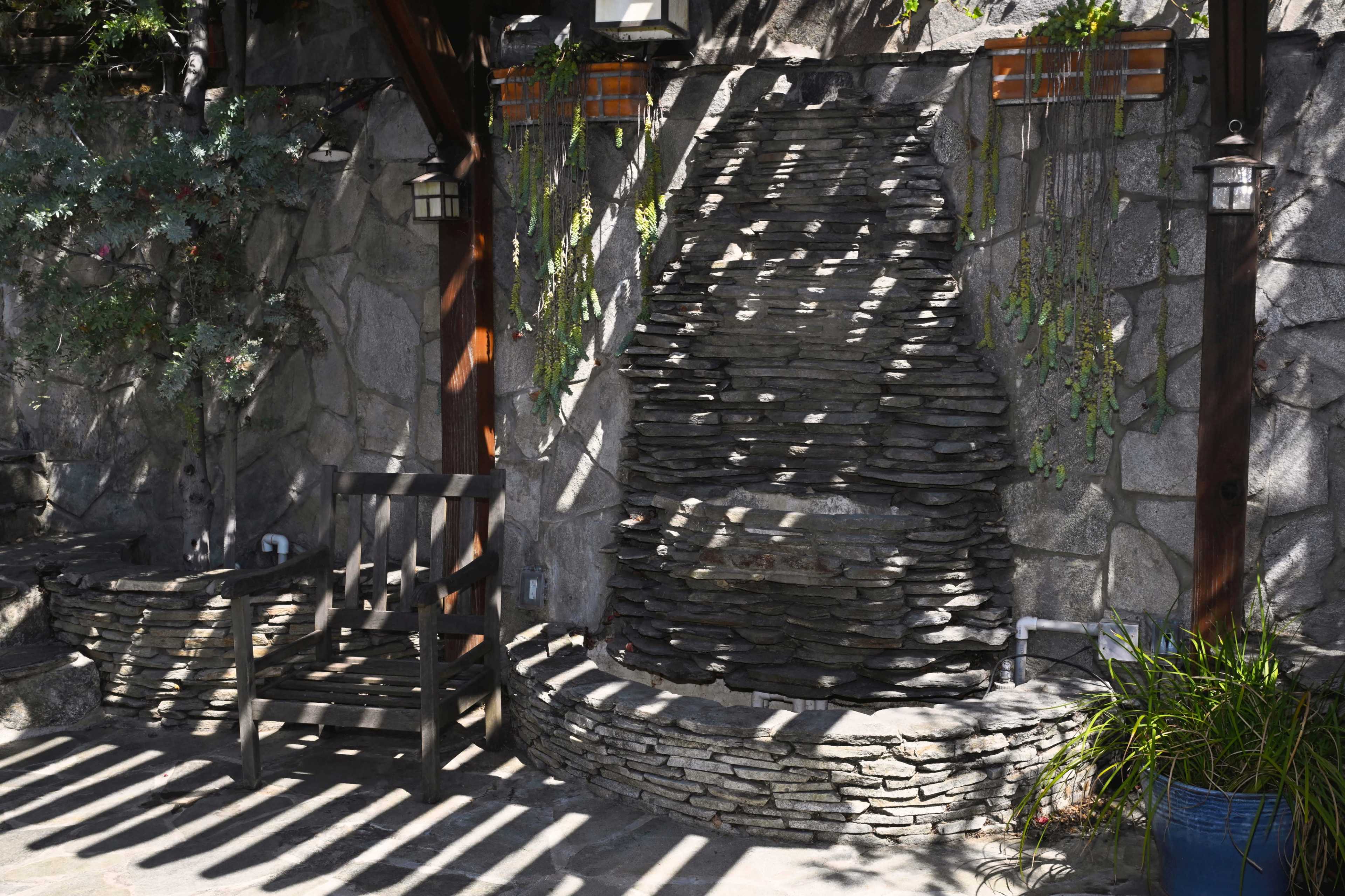 The image features a stone wall fountain surrounded by a wooden bench and lush greenery in a shaded outdoor space.