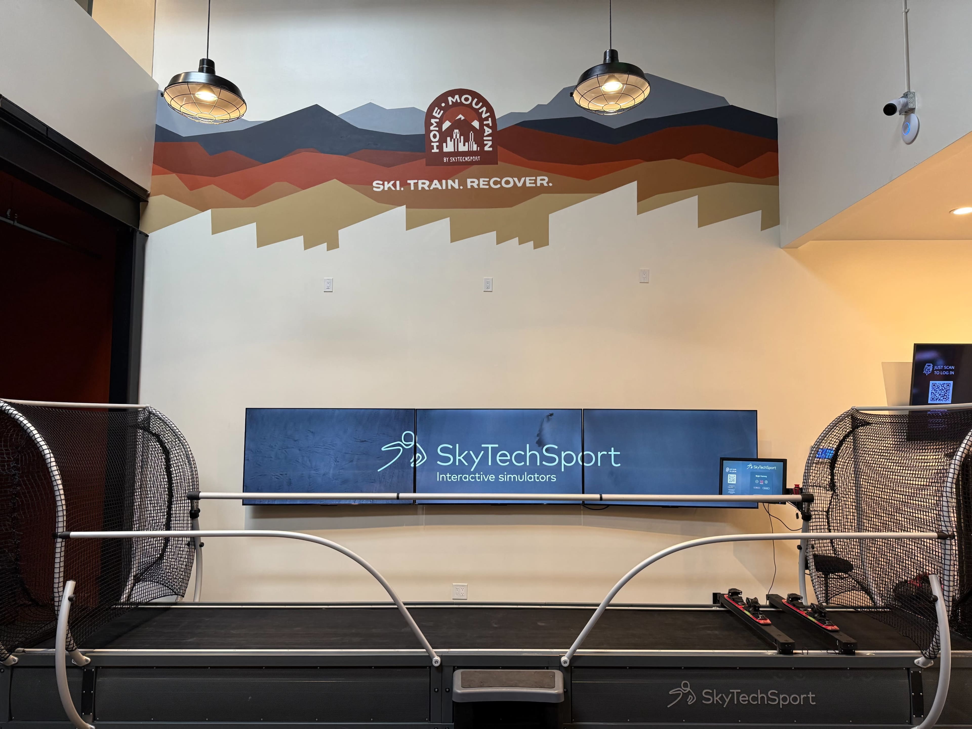 The image shows a ski training facility with a treadmill-like simulator and a large screen displaying the SkyTechSport logo and branding on the wall above.