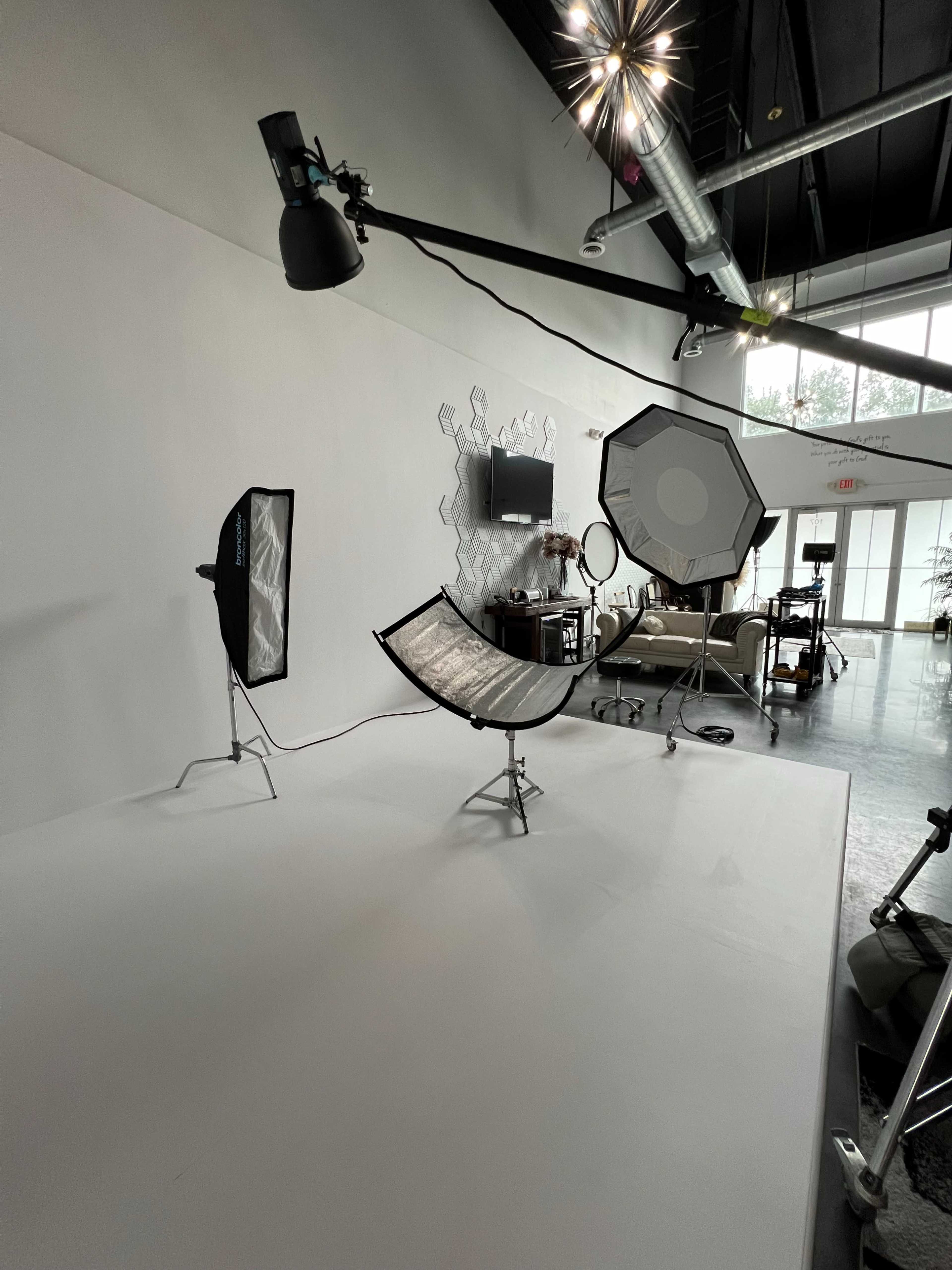 The image shows a photography studio setup featuring various lighting equipment and a simple backdrop, with a curved reflector and softbox placed on stands.