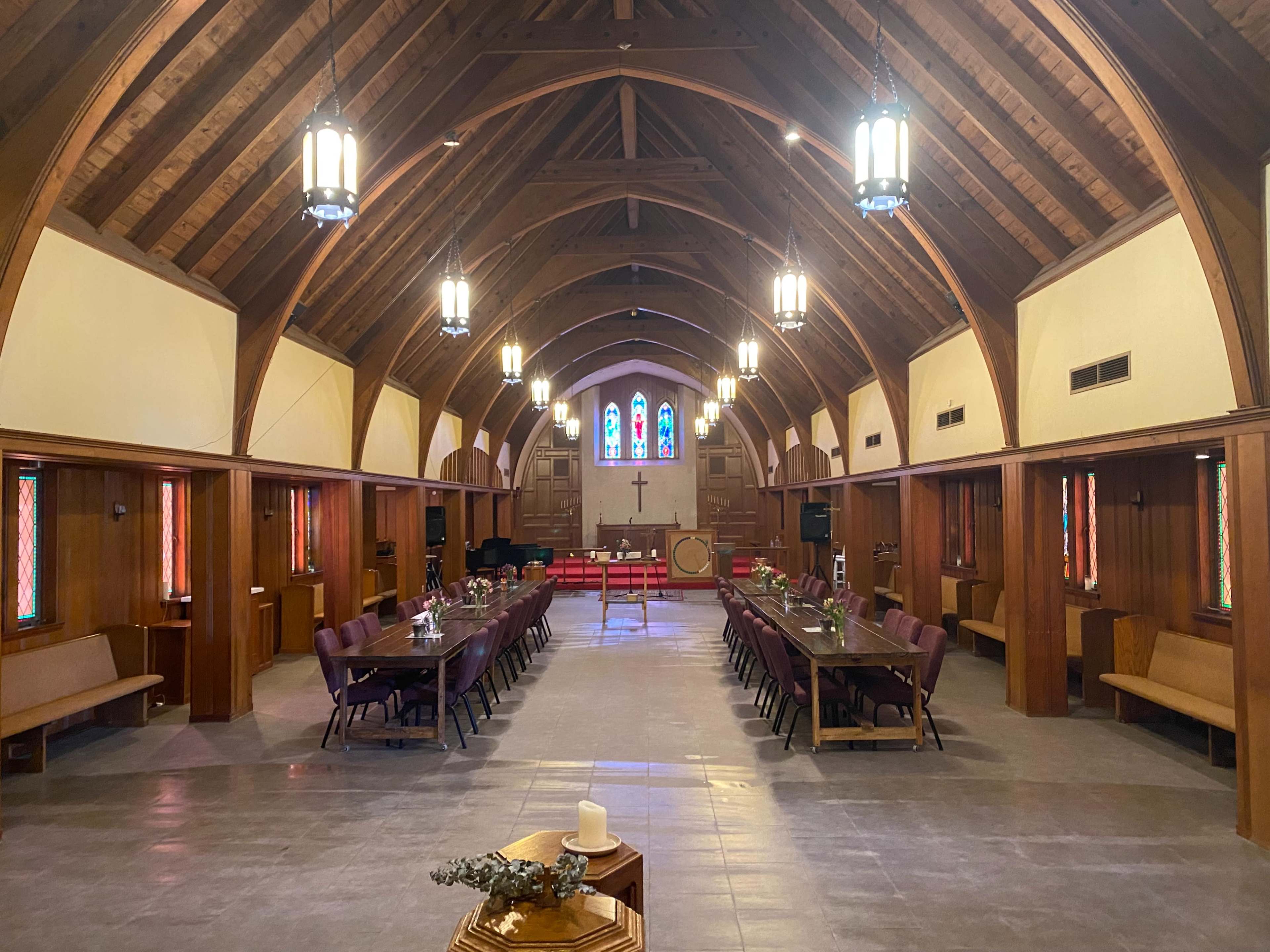The interior of a church features wooden beams, stained glass windows, and rows of tables set for a gathering.