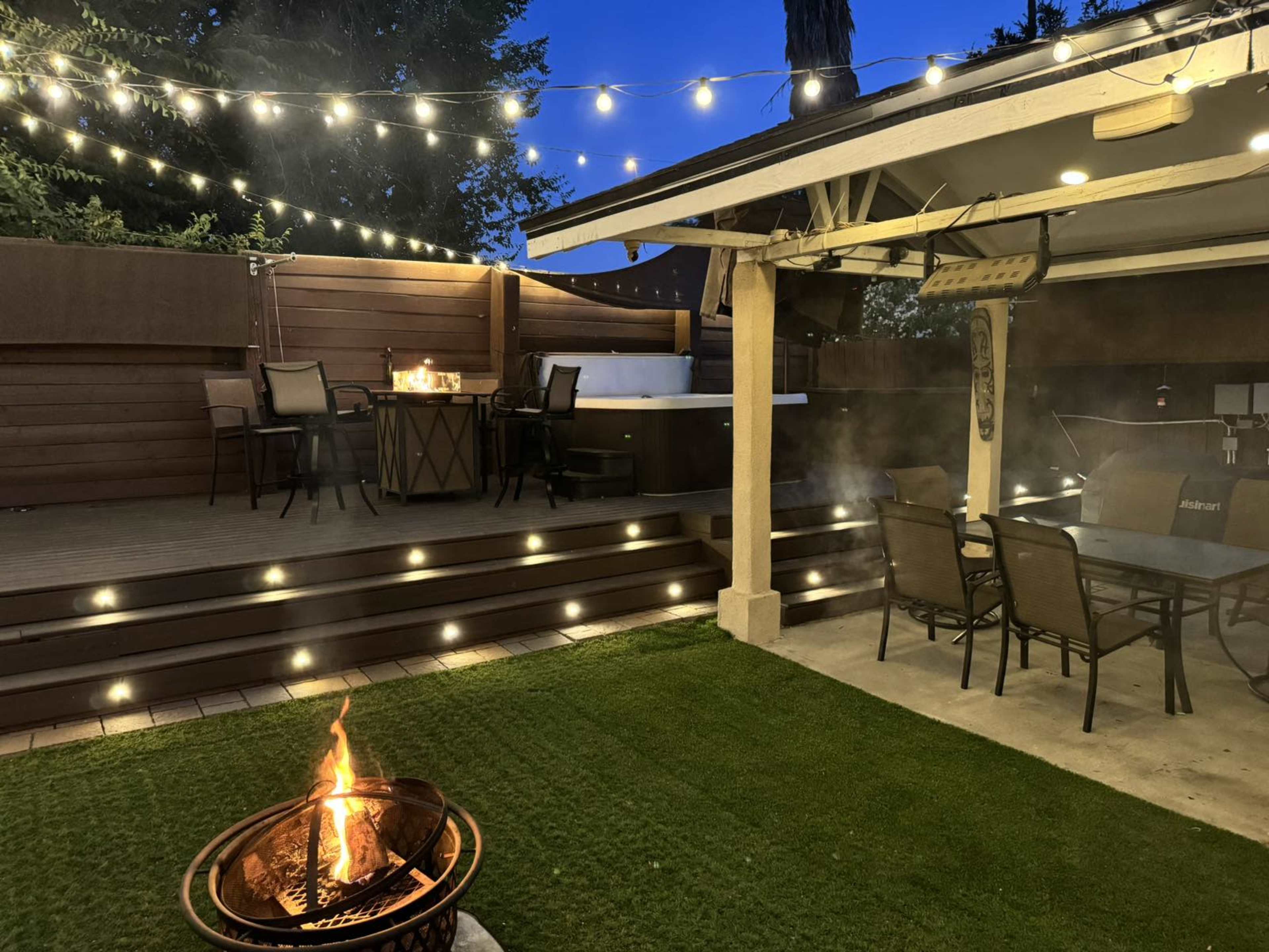 A backyard patio area with string lights, a fire pit in the foreground, a hot tub to the side, and a dining set on a wooden deck.