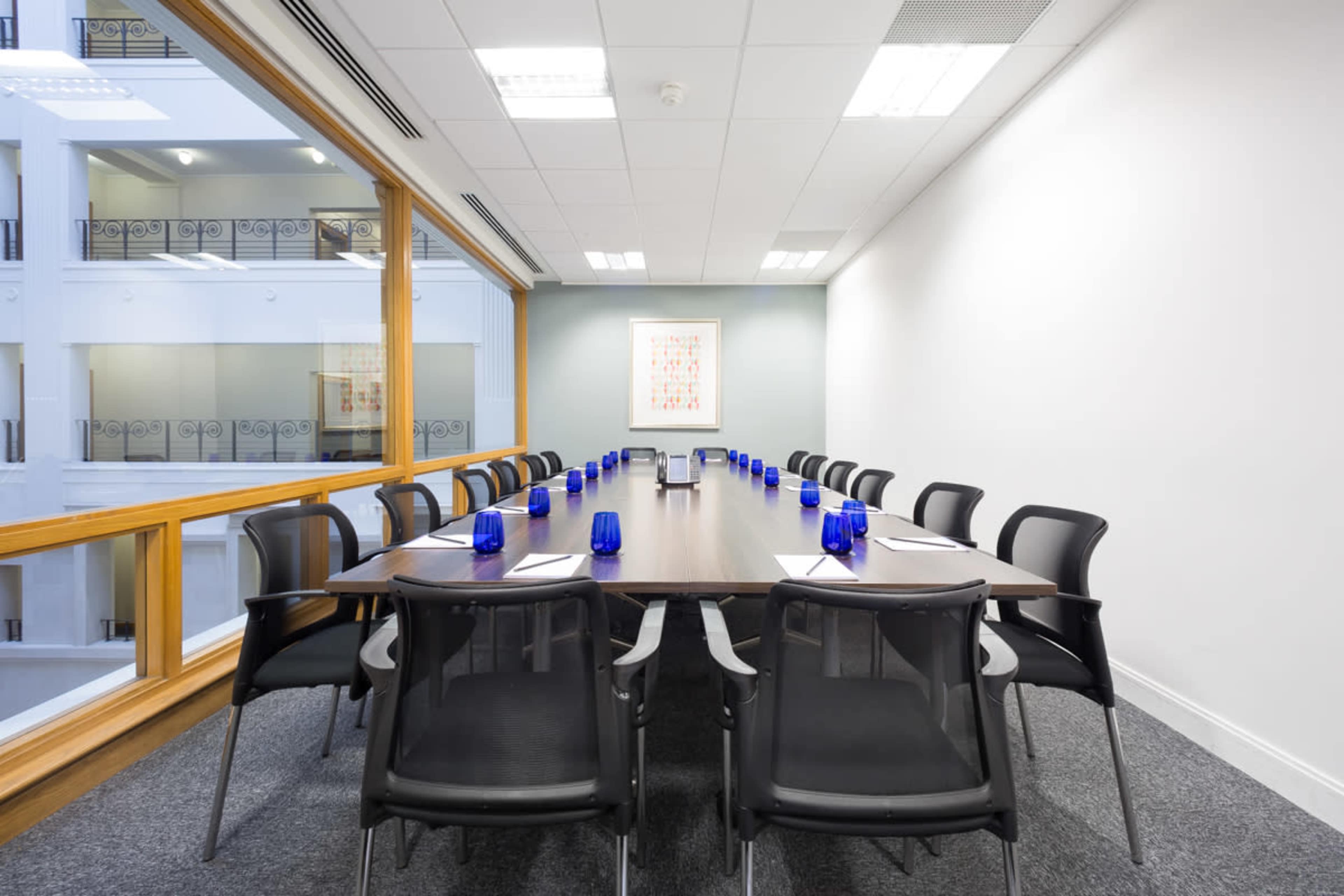 A conference room features a long table surrounded by black chairs, with blue glass candle holders and a large window overlooking an atrium.