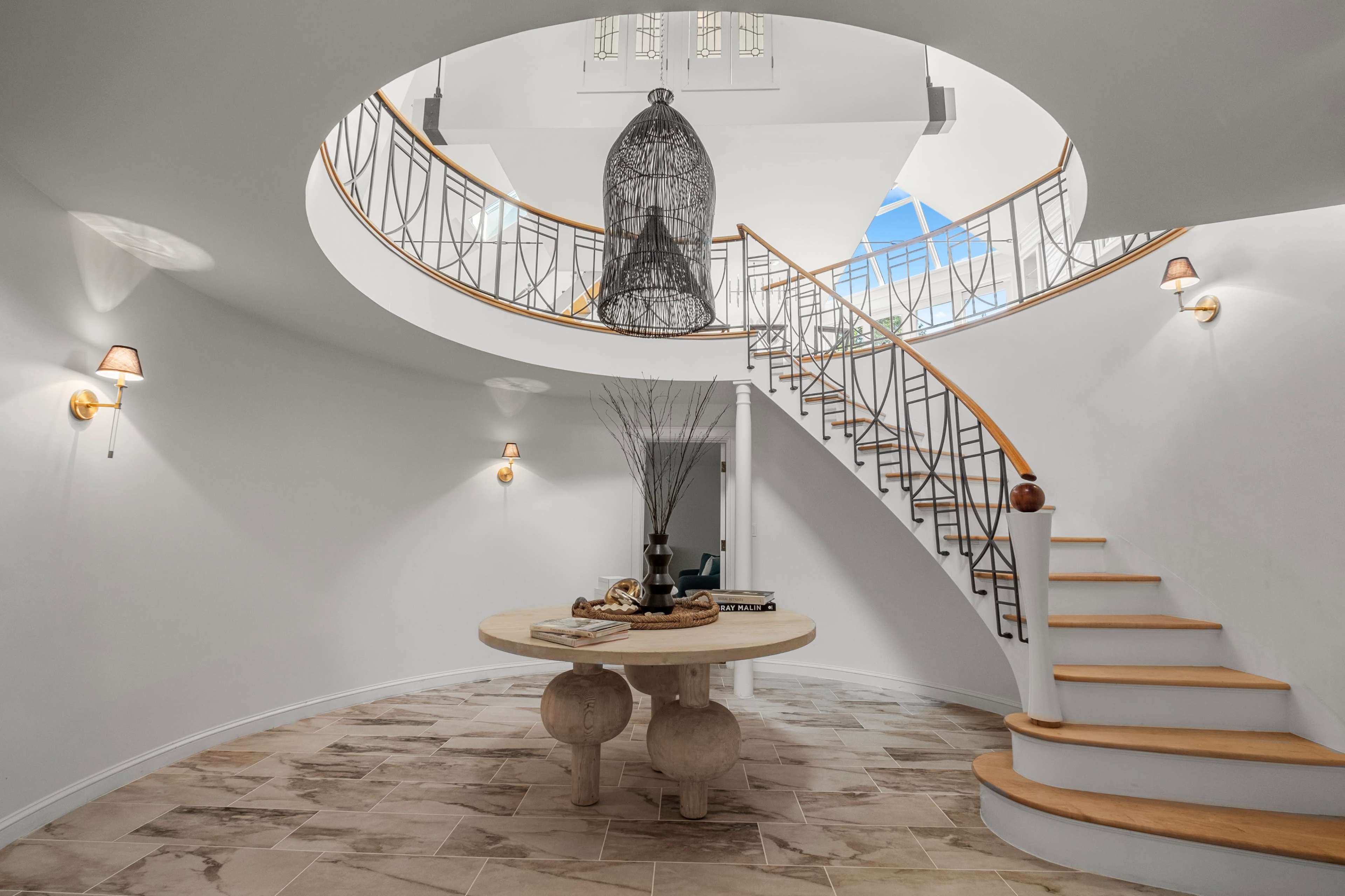 A circular entryway with a staircase ascending to a second level, featuring a large hanging light fixture and a round table in the center.