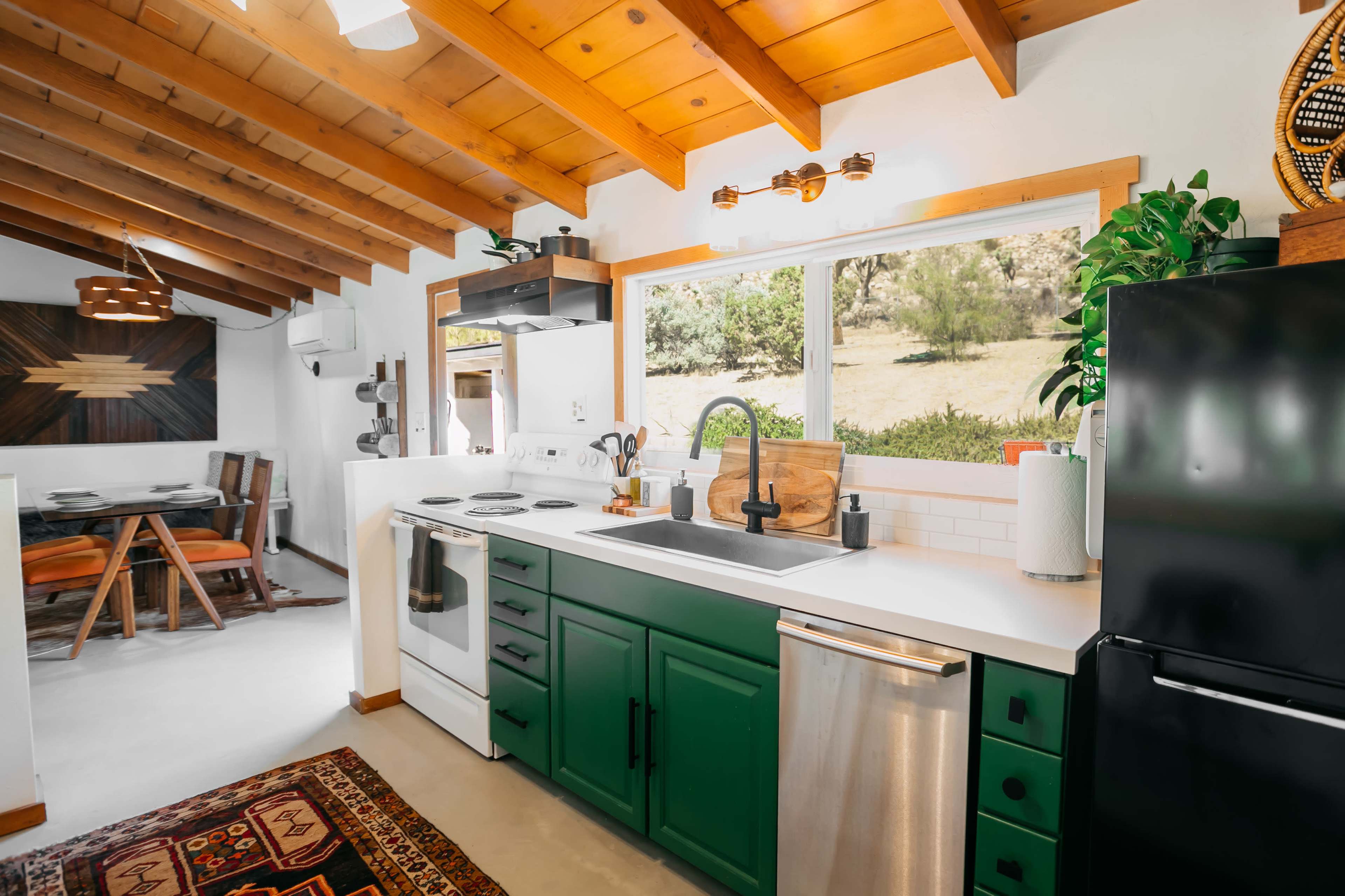 A well-designed kitchen with green cabinets, a white countertop, and a window overlooking a natural landscape.