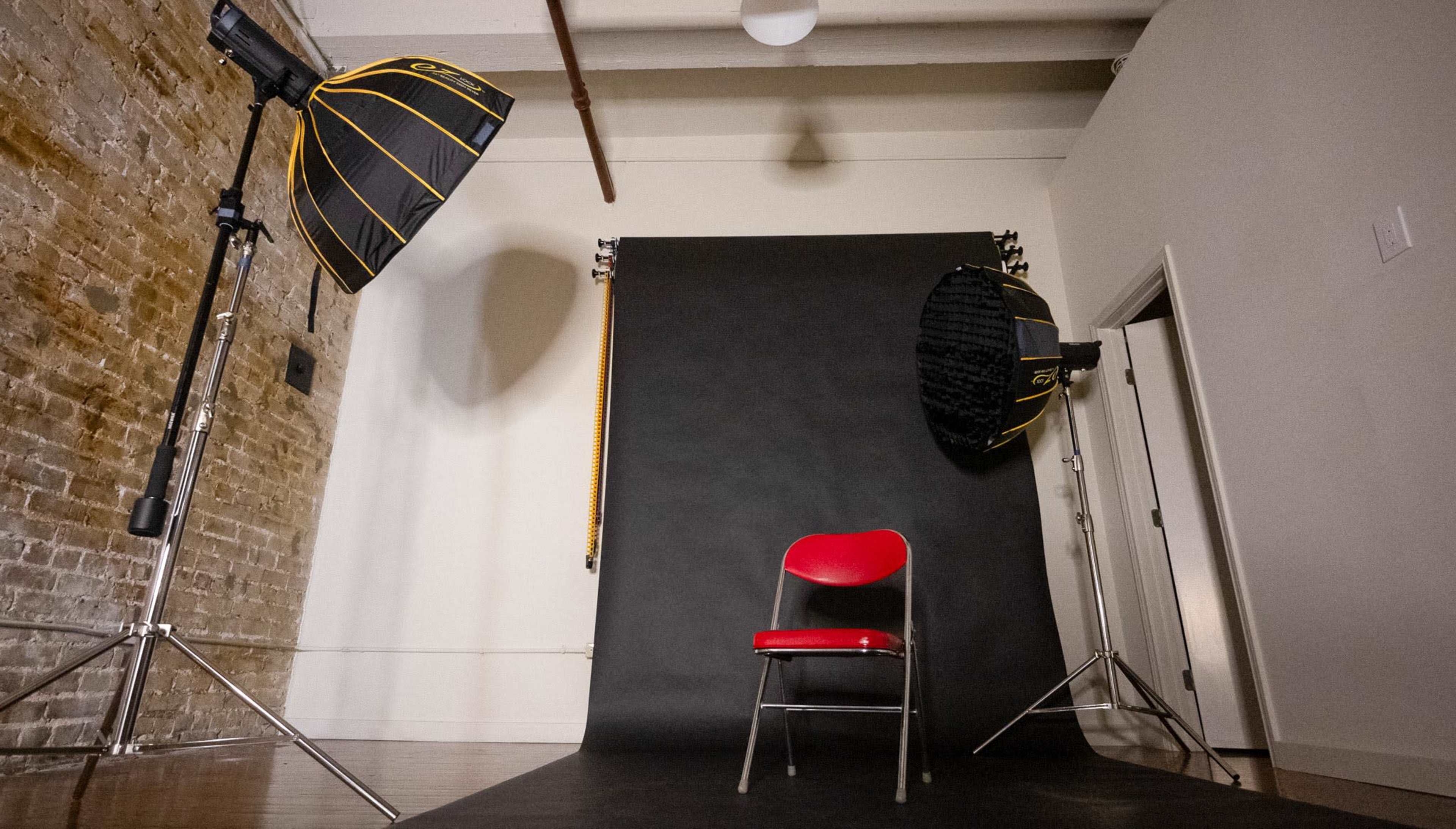 A red chair is positioned on a black backdrop in a photography studio with two lighting setups on either side.