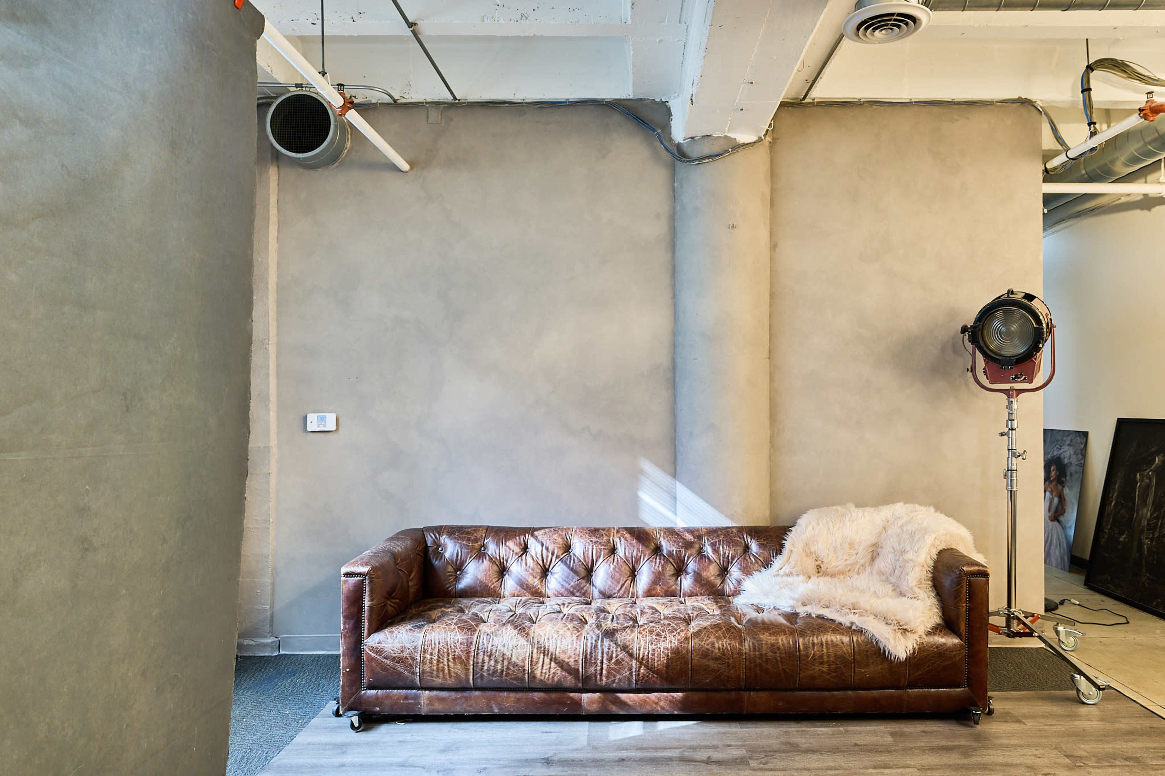 The image features a brown leather sofa against a plain, light gray wall with a vintage studio light positioned nearby.
