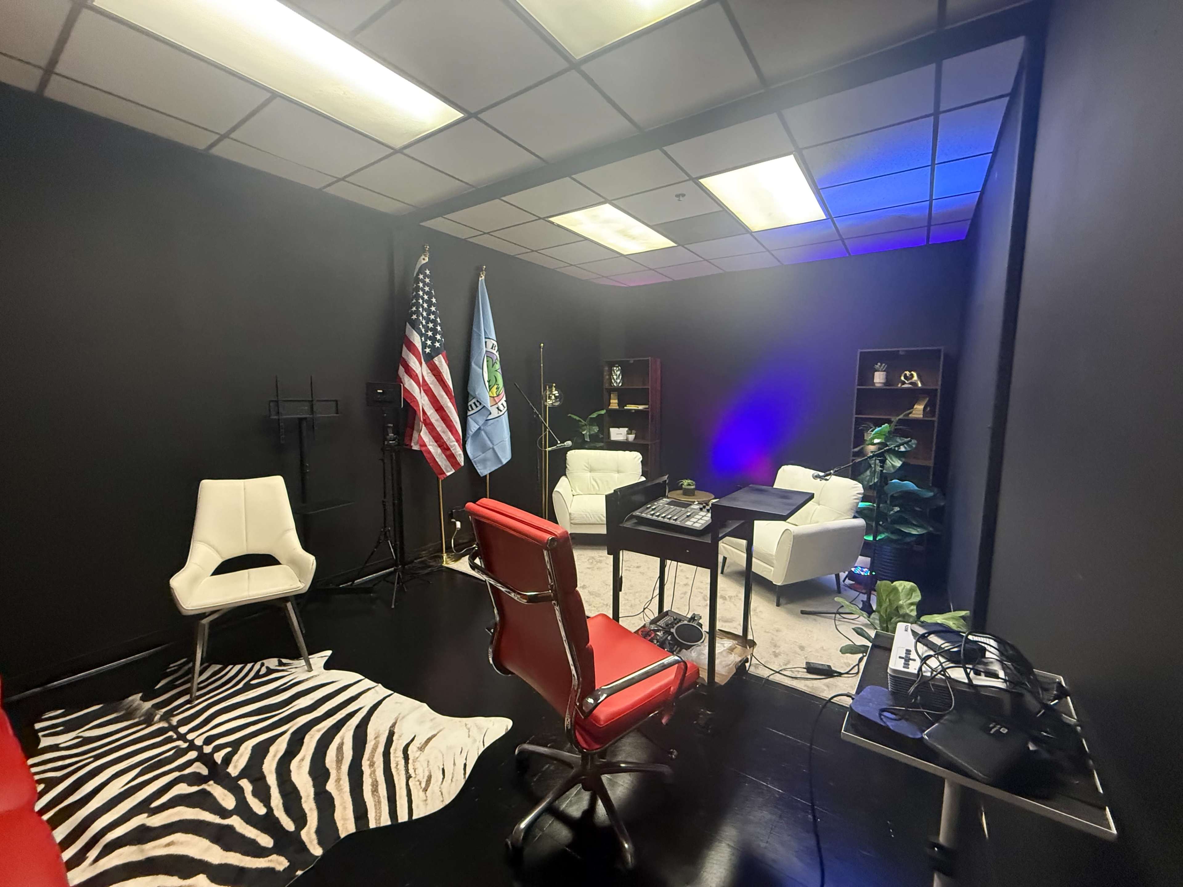 The room features a black wall with two white chairs, a red office chair, and a desk with equipment, accompanied by an American flag and a state flag.