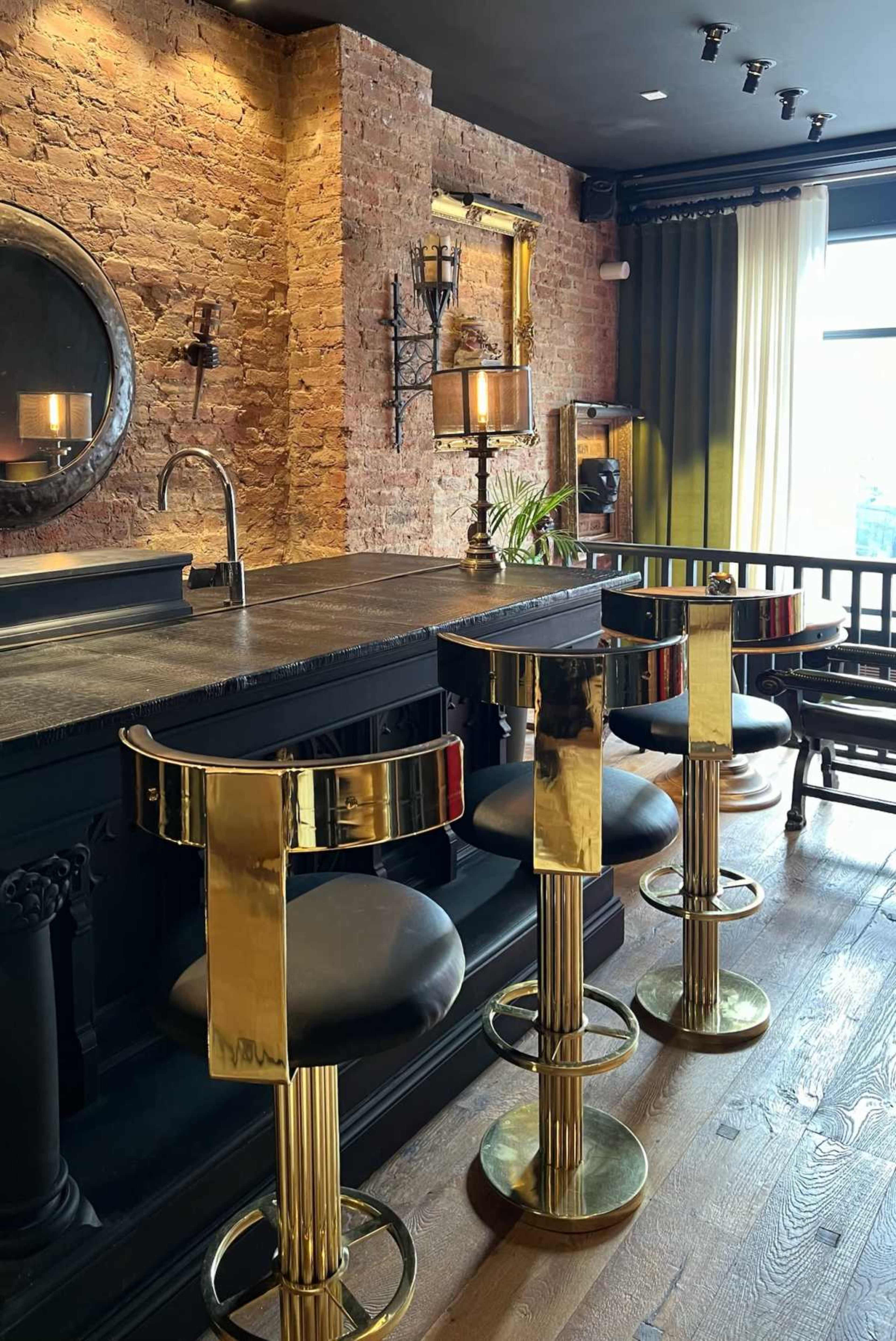 The image shows a modern bar area with a black countertop, gold-accented stools, exposed brick walls, and large windows providing natural light.