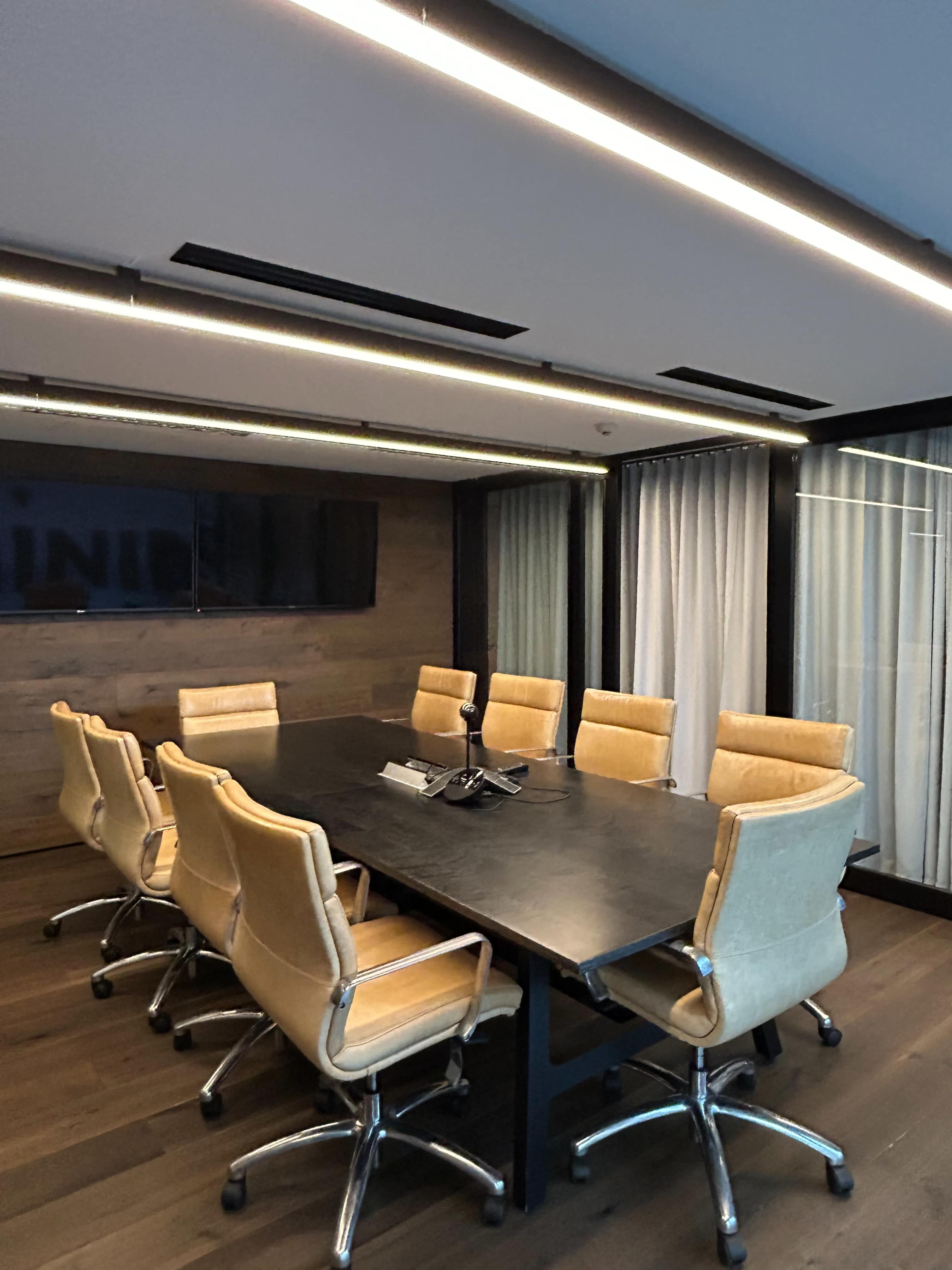 The image shows a modern conference room with a large black table surrounded by eight tan leather chairs and a wall-mounted screen.